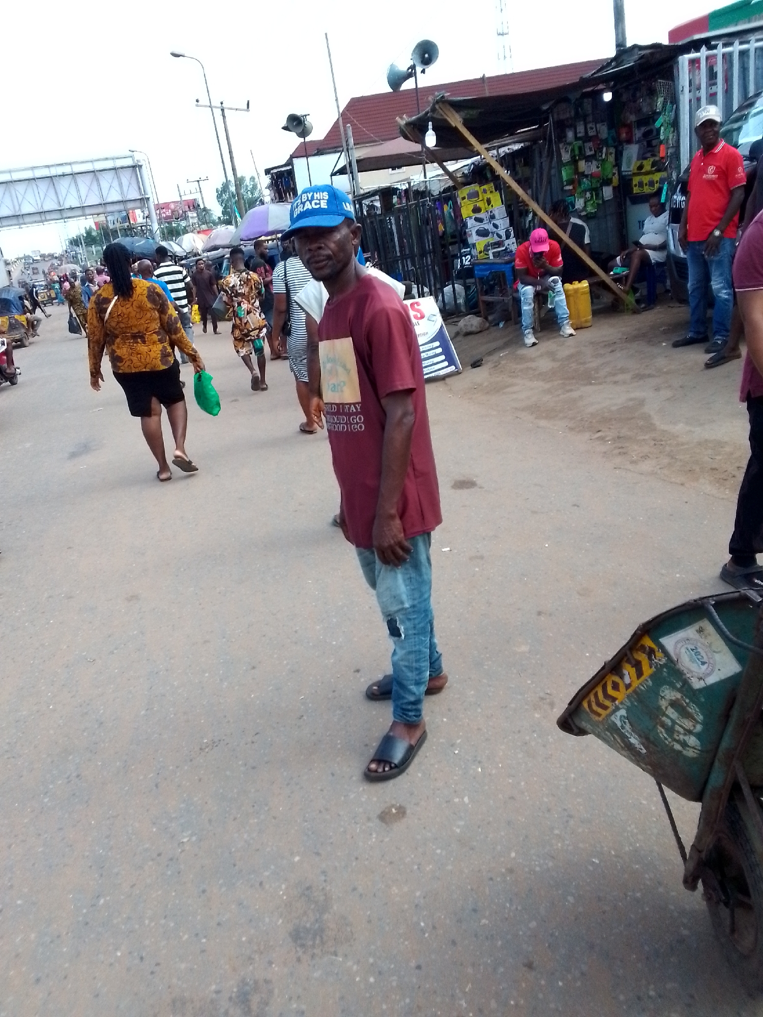 The image depicts a busy street scene in Onitsha, Nigeria. The foreground is dominated by a man in a maroon t-shirt and ripped blue jeans, wearing a blue baseball cap that says "BY HIS GRACE". He is standing on a paved road and looking towards the camera. To his right, a green wheelbarrow with yellow and black markings and stickers, one showing "2024", is partially visible. In the background, a diverse crowd of people are walking and browsing at stalls. One stall appears to sell electronics and accessories. The overall atmosphere is that of a typical market day in a Nigerian city, with a mix of commercial activity and pedestrian traffic.