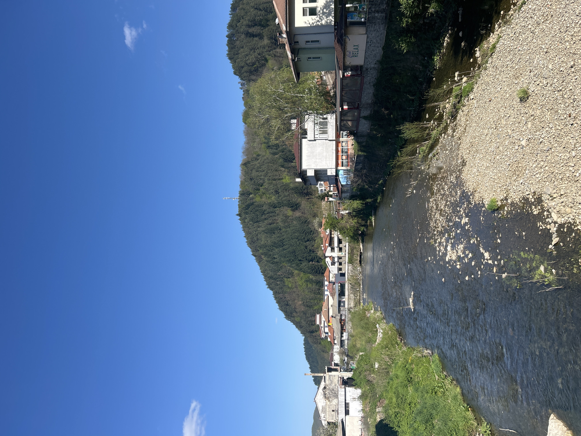 This image captures a serene landscape of a town situated alongside a river, nestled at the base of a forested mountain. The scene is bathed in bright daylight under a clear, azure sky with only a few wispy white clouds.
A river, likely shallow with its pebbly bed visible in many places, flows through the right and central parts of the frame. Its right bank features a wide expanse of light-colored pebbles and rocks, interspersed with small patches of green grass. The left bank is greener, with dense vegetation and trees.
Along the left bank, a series of multi-story buildings line the river. These structures vary in architectural style and color, suggesting a mix of residential and commercial properties. One prominent building displays a visible sign that reads "Cafe RELAX". Further along the river, towards the foreground, an older, possibly industrial building with a tall chimney can be seen.
Behind the buildings, a large hill or mountain rises steeply, densely covered by a dark green, mature forest, suggesting a natural and rugged environment.
The overall impression is one of tranquility and natural beauty, characteristic of a small town integrated with its natural surroundings. The bright sunlight indicates clear weather, likely late morning or early afternoon. The location context provided is Zlatograd, Bulgaria.