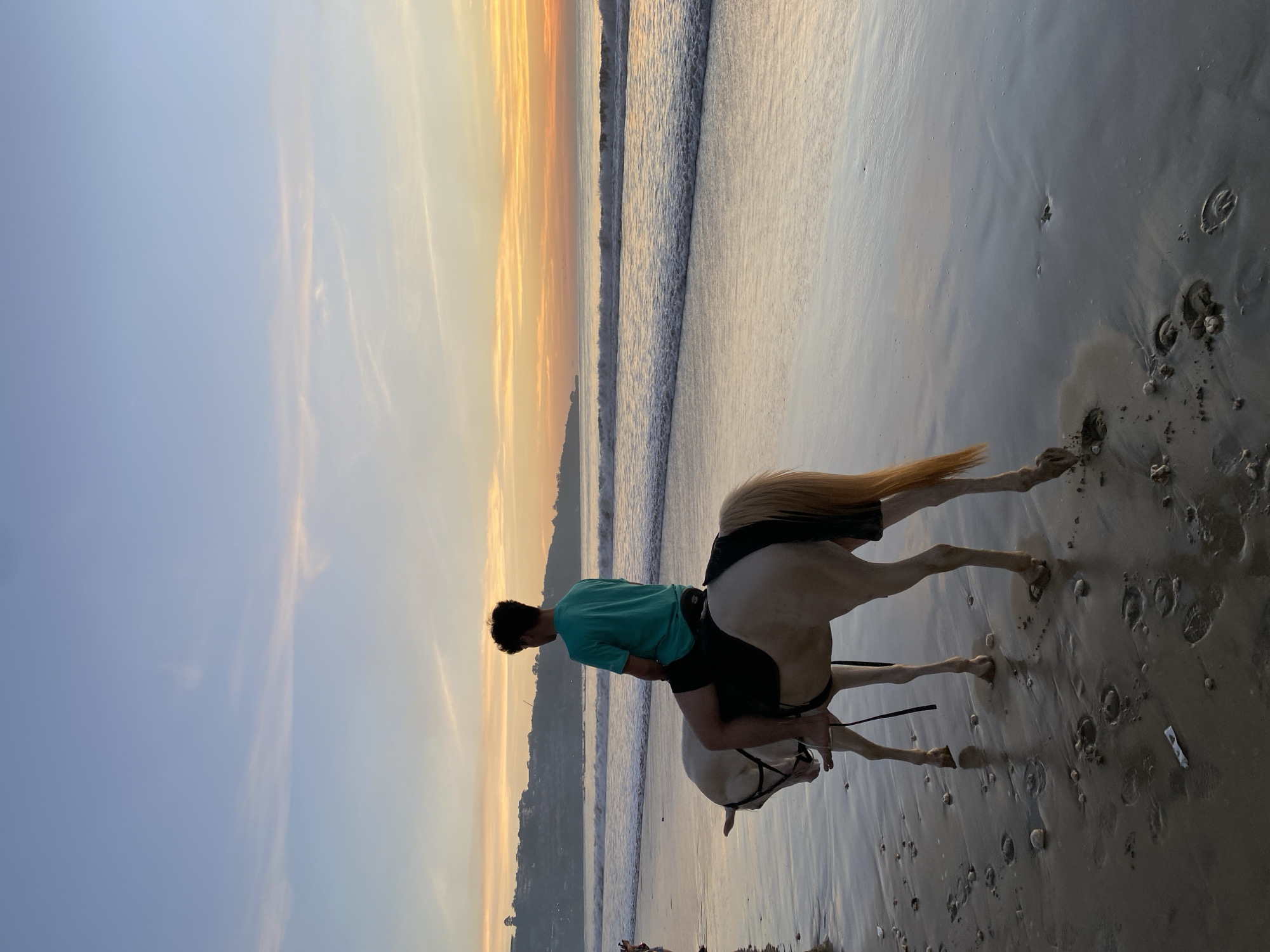 A man in a green t-shirt rides a pale horse on a beach in Kuta Selatan, Indonesia, at sunset. Waves break on the shore as the horse walks along the wet sand, leaving hoofprints. The sky shows shades of blue, orange, and yellow.
