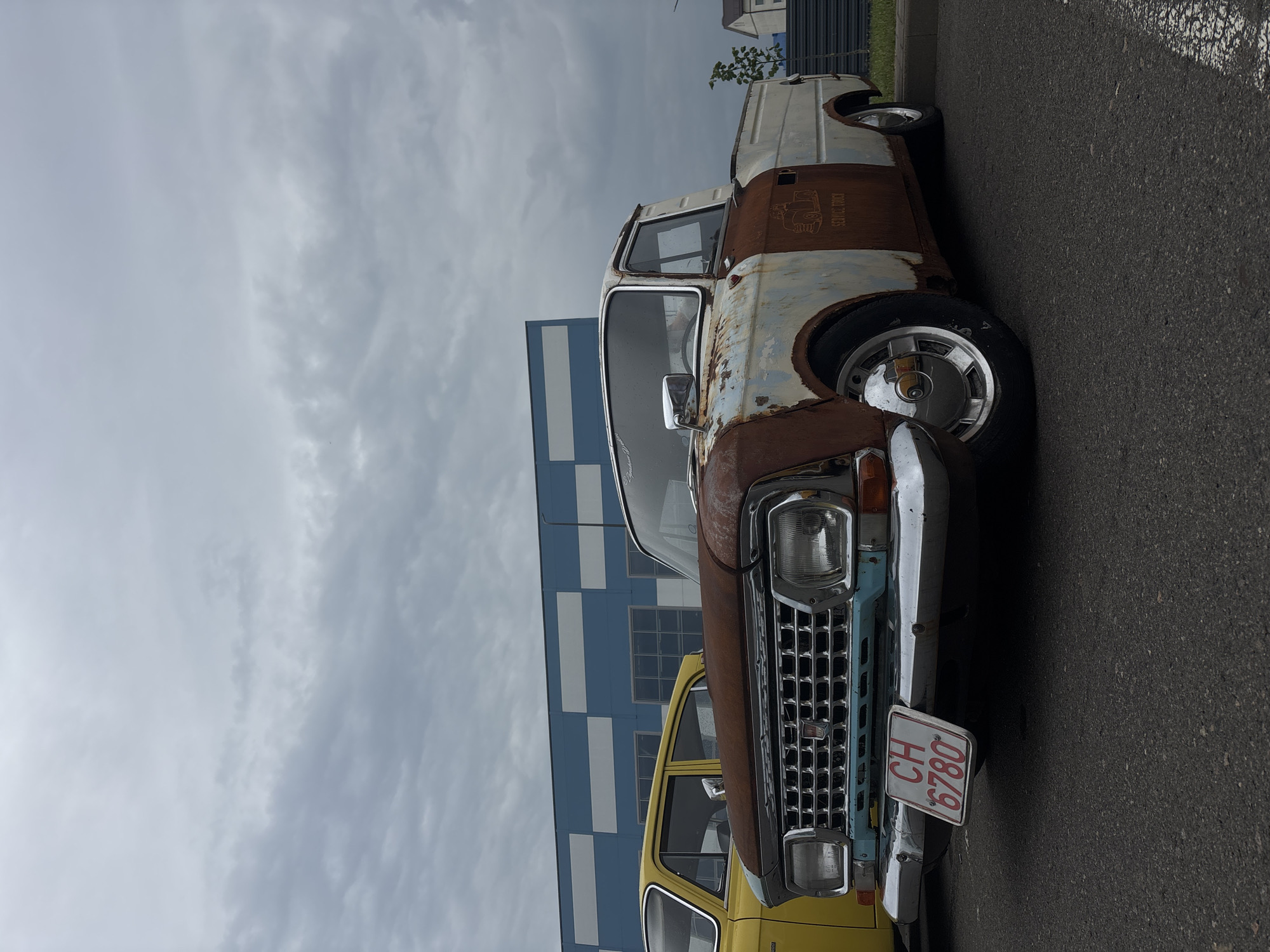 In Uzda, Belarus, a heavily rusted white pickup truck with a white cab and a light blue grille is parked on an asphalt surface. The truck has a dark metal fender and a chrome bumper with a license plate that reads "CH 6780". A yellow sedan is parked to the left of the truck. In the background, a building with blue and white checkered patterns is visible. The sky is overcast with gray clouds.