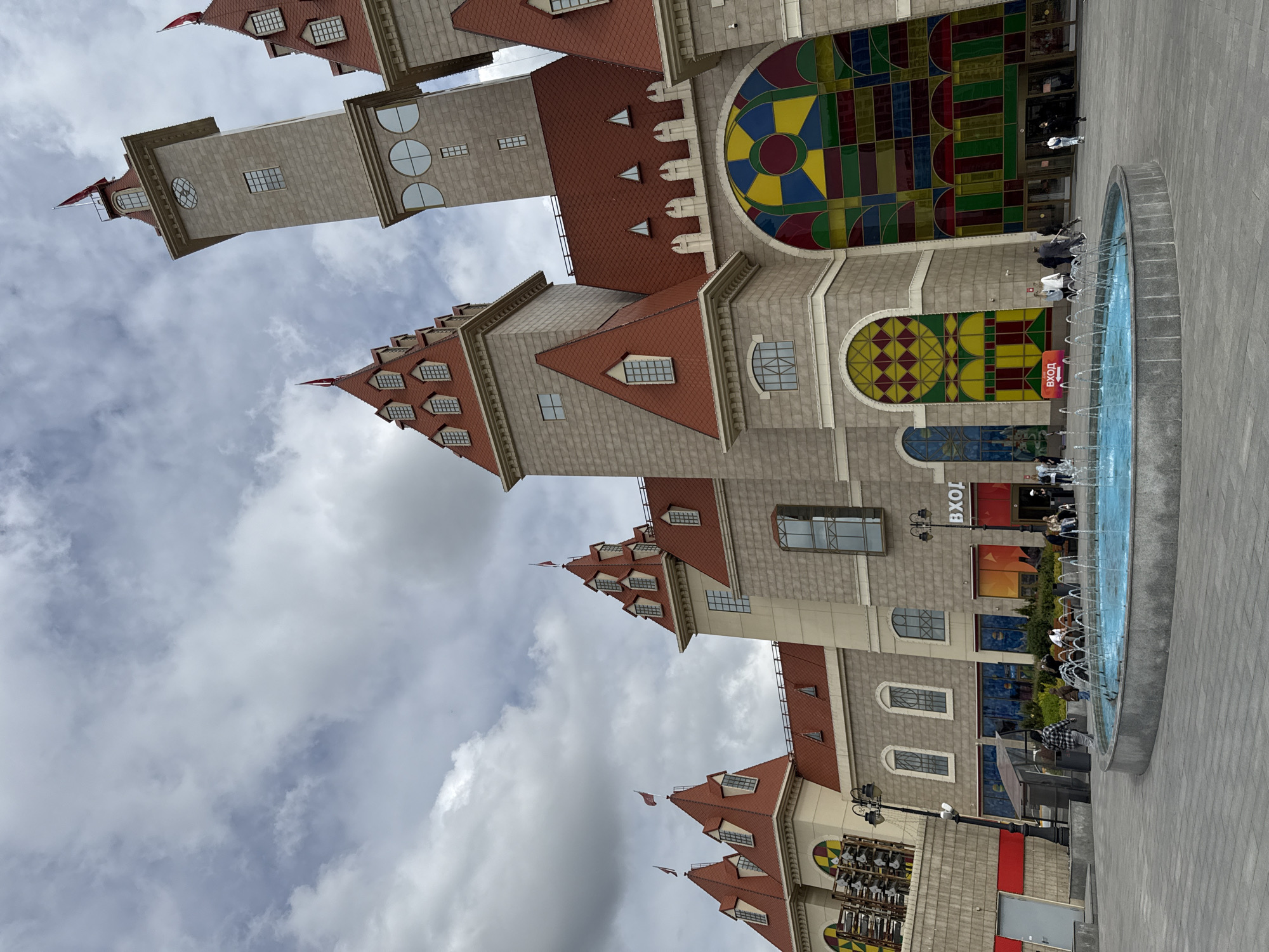 A grand, multi-towered building complex with architecture reminiscent of a fairytale castle dominates the scene under an overcast, cloudy sky. The structure features light grey stone or stucco facades and distinctive red-tiled roofs with numerous dormer windows and small turrets, many adorned with small red flags.
Large sections of the building's exterior boast striking, colorful stained-glass-like windows, displaying geometric patterns in shades of red, yellow, blue, and green. Various other window shapes, including arched, circular, and rectangular, are also visible across the different levels of the building.
In the foreground, a circular fountain with multiple upward-spraying jets of water is set into a paved plaza. Several people are scattered throughout the outdoor area, some walking near the building's entrances, and others seated or standing around the fountain or along the building's edge. They appear to be casually observing their surroundings or engaging in leisure activities.
A prominent red sign with white Cyrillic text, "ВХОД" (VKHOD), meaning "ENTRANCE," is visible on one of the building's facades, indicating an entry point. The scene depicts a daytime setting with diffused light from the cloudy sky. The architectural style suggests a modern entertainment or commercial complex with a whimsical theme, located in Moscow, Russia.