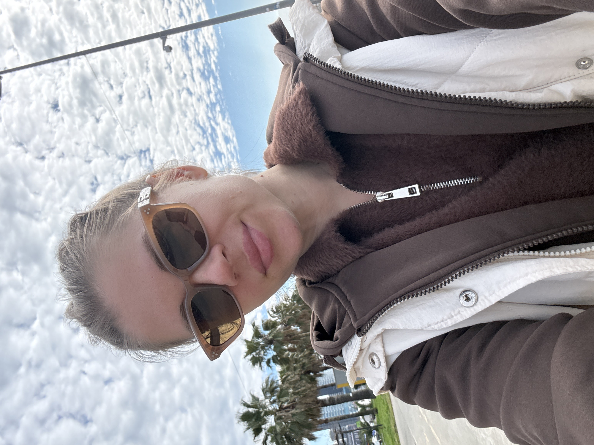 The image is a tilted, eye-level selfie featuring a person outdoors on a bright day in Batumi, Georgia. The subject is a light-skinned individual with light-colored hair, wearing brown-framed sunglasses that cover their eyes. Their lips are slightly parted. They are dressed in a brown hooded jacket or sweatshirt, which appears to have a soft, furry brown lining visible around the collar and chest area. A metallic zipper is prominent on the jacket. Underneath, a white quilted layer is visible, also featuring a zipper and snap buttons.
The background primarily consists of a bright blue sky filled with numerous fluffy white clouds, indicating clear weather. Towards the lower right, several green palm trees are visible, suggesting a warm, possibly coastal environment consistent with Batumi. Hints of urban structures or buildings can be seen beyond the palm trees. A dark horizontal bar, possibly part of a lamppost or overhead structure, crosses the upper part of the sky. The overall impression is a casual outdoor shot, capturing a moment under sunny skies. No discernible text is visible in the image.