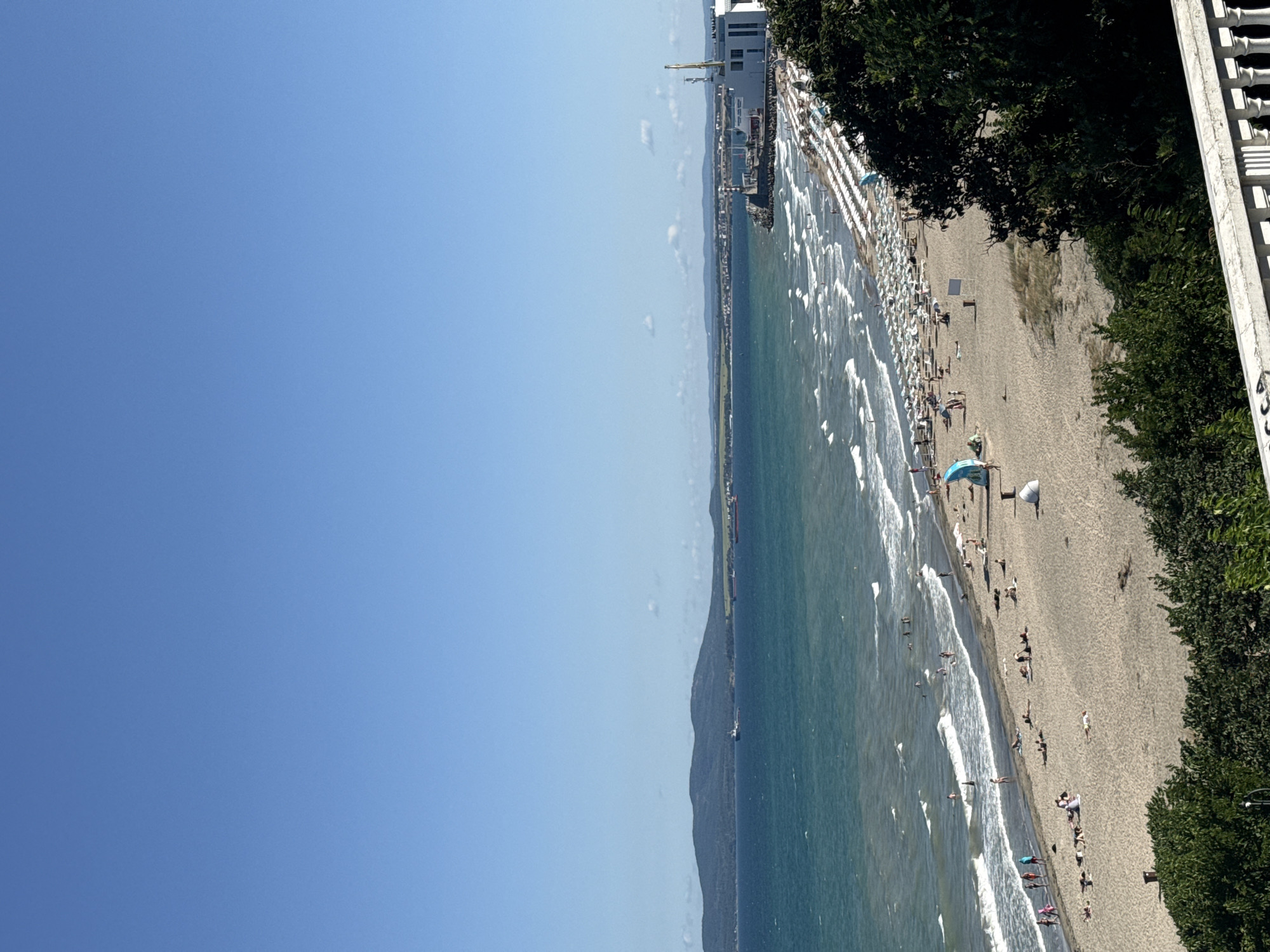 This is a wide-angle, high-angle photograph of a crowded beach on the Black Sea coast, likely in the city of Burgas, Bulgaria. The clear blue sky dominates the upper half of the image. Below the sky, a vast expanse of turquoise water stretches to the horizon, with gentle waves breaking on the shore.
The beach itself is a wide strip of light-colored sand, densely populated with beachgoers. Numerous white beach umbrellas and loungers are arranged in neat rows along the shoreline, indicating a developed resort area. People are seen sunbathing, walking along the beach, and swimming in the sea. Some individuals are gathered in small groups, interacting socially. In the distance, along the coastline, a pier or harbor area is visible with several buildings and a crane. A few ships are present in the sea, including one closer to the shore and another further out towards the horizon.
The foreground is partially obscured by lush green foliage and a white balustrade, suggesting the photo was taken from an elevated vantage point, possibly a balcony or a viewpoint overlooking the beach. The overall scene depicts a lively summer day at a popular seaside destination.