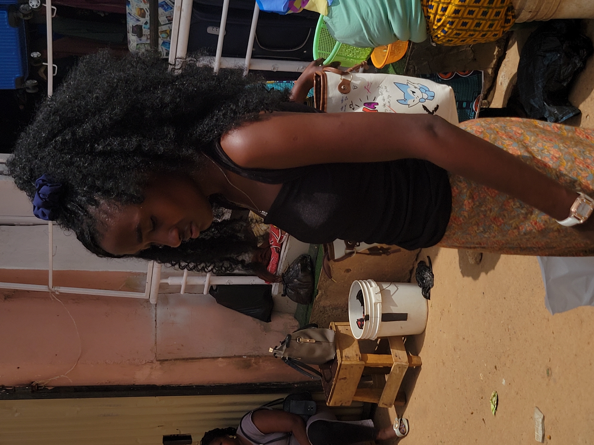 A woman with dark, curly hair stands outside a building with a peach-colored exterior wall, presumably in the city of Jos, Nigeria. She wears a black tank top, a patterned skirt, and a white watch. Her right hand is extended, holding a white handbag with colorful designs. Behind her, there are various objects including luggage, plastic baskets, and what appears to be laundry on a drying rack. To her right, there's a white bucket on a wooden stand and a bag nearby. In the lower right corner, there is a white cloth and dirt. A person is on the floor towards the bottom of the image.