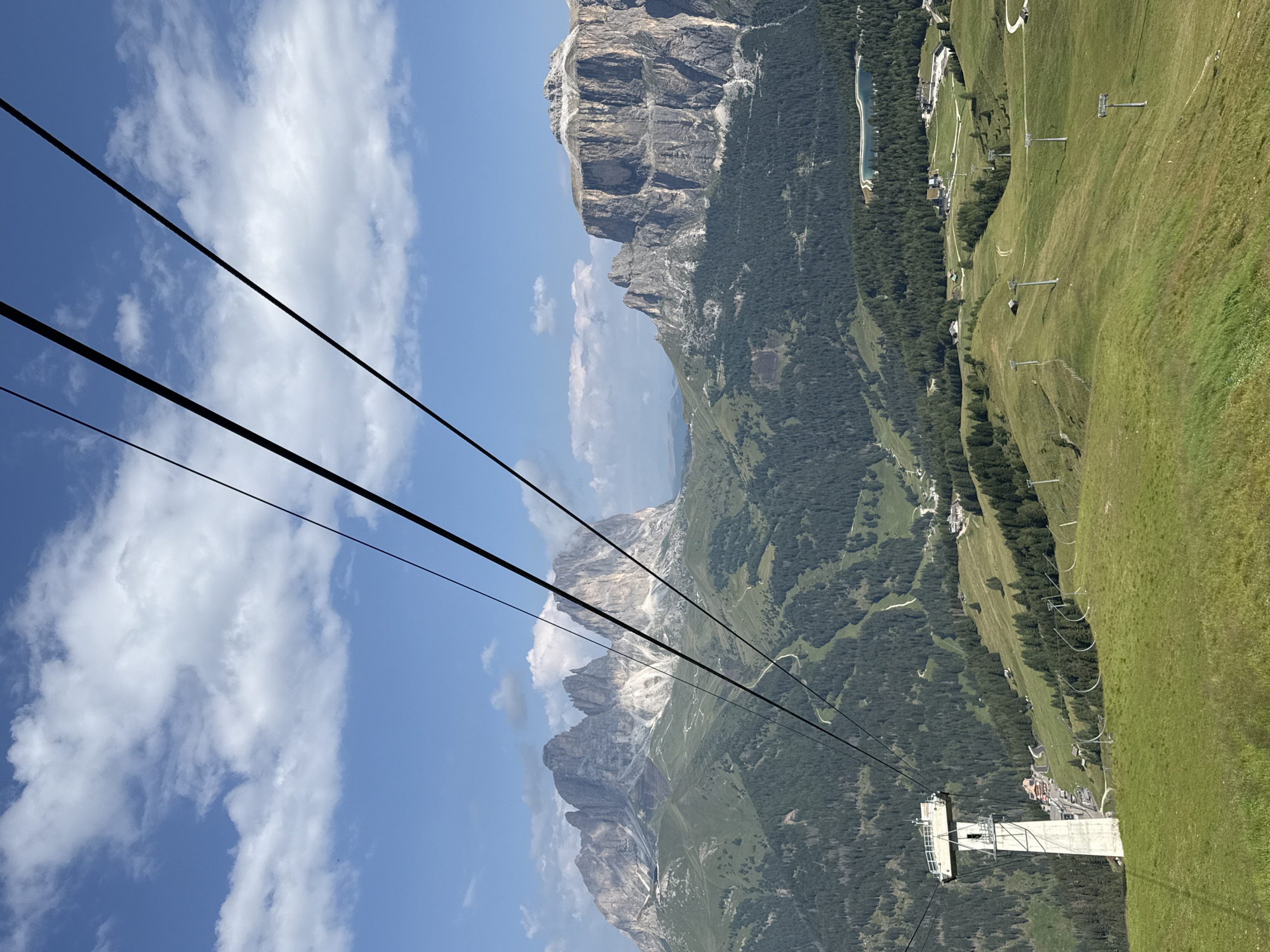 This is a wide-angle, eye-level, outdoor, daytime photograph taken from a cable car in the Dolomite Mountains, near Canazei, Italy. The image features a dramatic mountain landscape with rugged peaks in the background and a lush green valley in the foreground. Several ski lift towers and cables are visible, indicating the area is a popular ski resort. The sky is a bright blue with scattered white clouds.
In the foreground, a concrete ski lift tower stands on a grassy slope. The tower is tall and utilitarian, with metal scaffolding and lights. Multiple black cables stretch diagonally across the frame, suggesting the viewer is currently in transit via the cable car.
The mid-ground is characterized by rolling green hills dotted with dark green pine forests. Winding paths and roads are visible throughout the landscape, leading to scattered buildings and structures nestled within the valley. A small, serene lake with a distinctive crescent shape is visible in the distance, reflecting the sky.
The background is dominated by majestic, jagged mountain peaks, their rocky faces illuminated by the sun. Some of these peaks show patches of snow. The scale of the mountains is impressive, emphasizing the grandeur of the natural environment.
There are no people visible in the photograph. The overall impression is one of breathtaking natural beauty and a well-developed mountain resort.