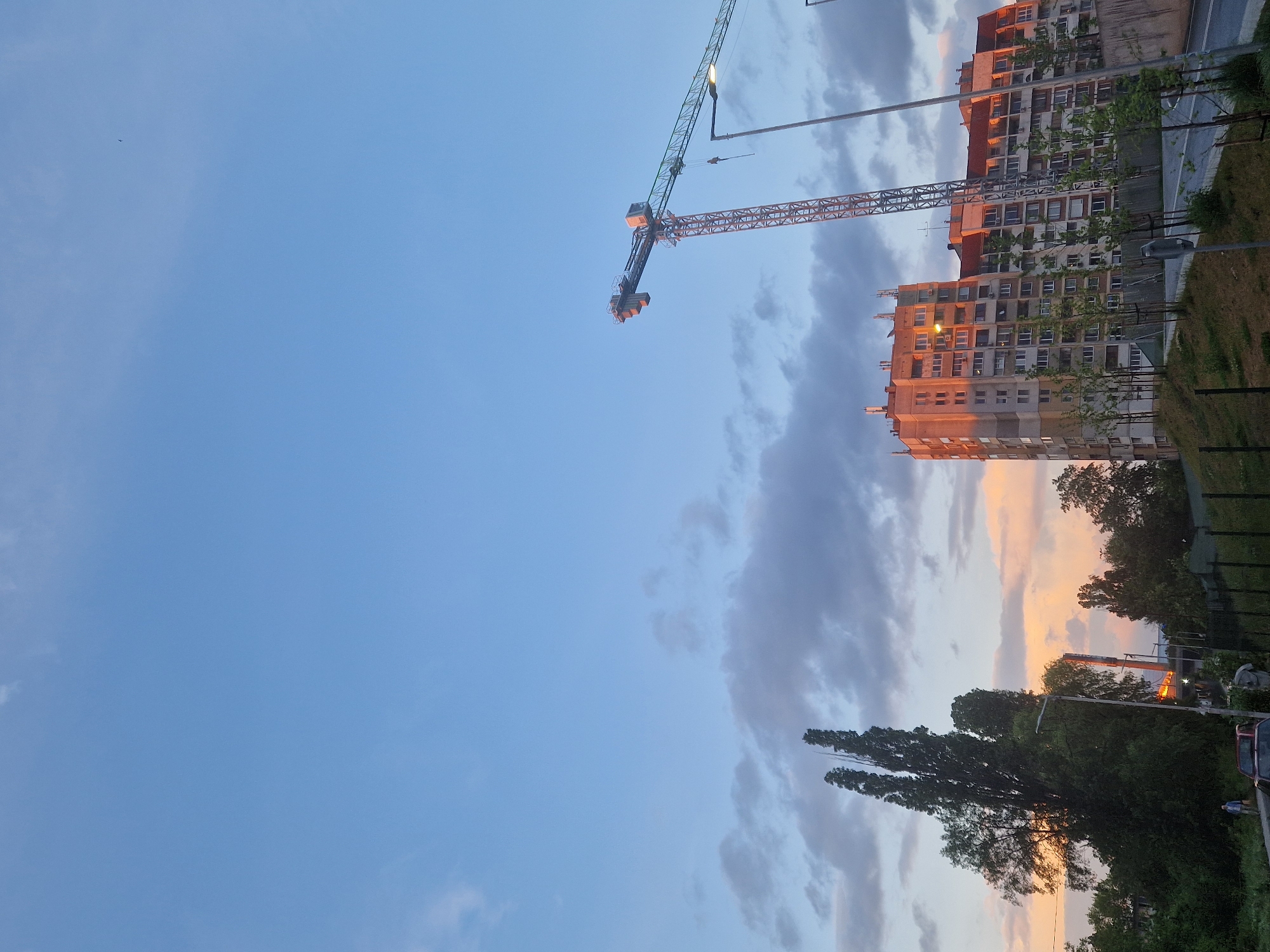 The image depicts a cityscape at dusk, possibly during twilight hours. In the center of the frame, a large construction crane is visible against a partly cloudy sky. A group of tall apartment buildings are located to the right of the crane, illuminated by a warm, orange light, likely from the setting sun. Trees are present in the lower right corner, partially obscuring some of the buildings. In the background, a portion of a street and sidewalk can be seen adjacent to a grassy area. The location of the city is unknown.