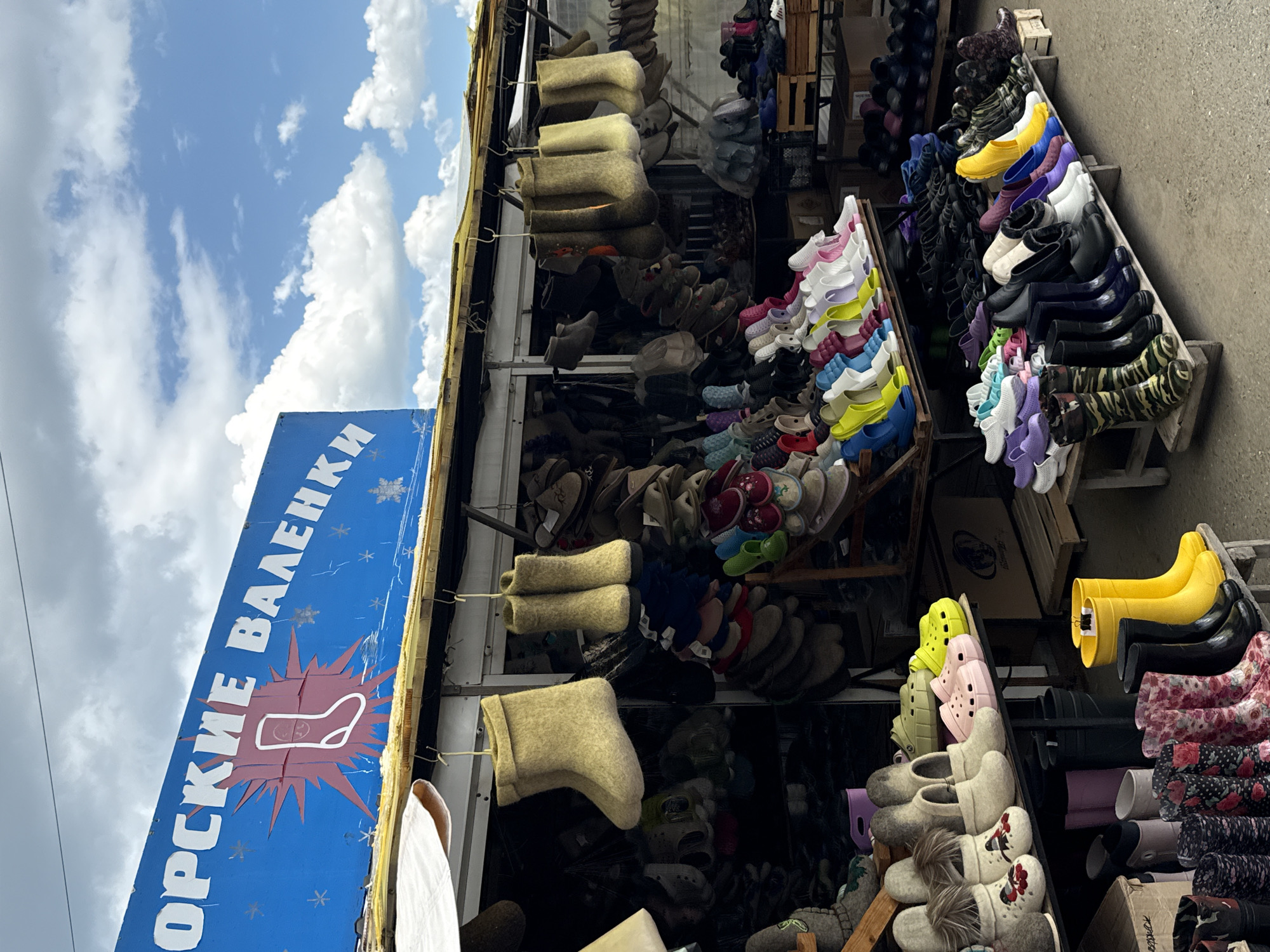 The image depicts an outdoor market stall in Izhevsk, Russia, specializing in felt boots and other footwear. A blue sign with white lettering, partially visible, reads "ОРСКИЕ ВАЛЕНКИ" which translates to "Orenburg Felt Boots," indicating the origin or style of the merchandise. The stall is filled with various types of footwear, including traditional felt boots (valenki) hanging from hooks, rubber boots in multiple colors, clogs, and other casual shoes. The products are displayed on shelves and hanging racks, suggesting a commercial establishment. The background features a cloudy sky, implying it's daytime. The overall scene captures the retail environment of a local market in Izhevsk.