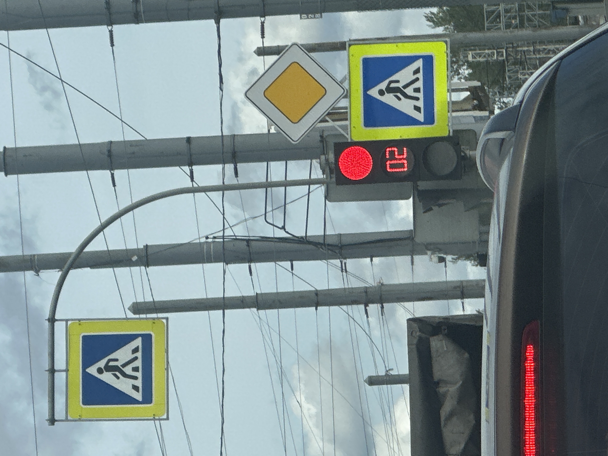 In Saint Petersburg, Russia, a traffic light displays a red signal and a countdown timer showing "02". Two pedestrian crossing signs are visible: one square with a blue background and a white triangle containing a black pedestrian symbol, outlined in yellow, and another identical sign below the traffic light. A diamond-shaped sign with a yellow center and a white border is mounted above the traffic light. The scene is framed by utility poles and wires, with a car's rear brake light illuminated in the foreground. The sky is overcast with clouds.