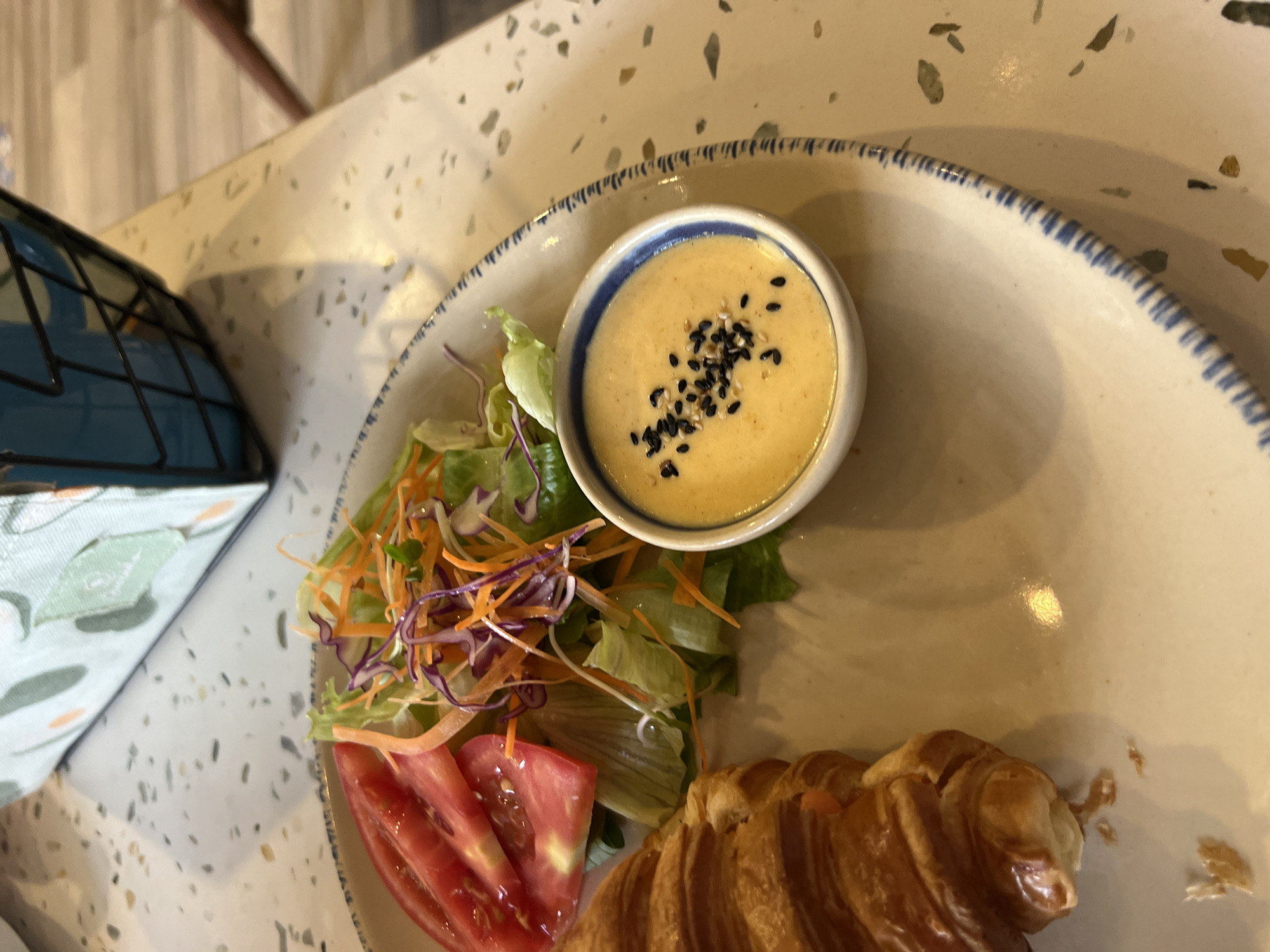A high-angle, close-up shot shows a partial view of a plate containing food. The plate is circular with a white base and a blue patterned rim. On the plate, there is a croissant, sliced tomatoes, a salad with shredded carrots and purple cabbage, and a small white bowl containing a yellow sauce speckled with black seeds. The table beneath the plate has a speckled beige and white surface. The scene does not contain identifiable people or actions, and the location is not discernable from the provided context.