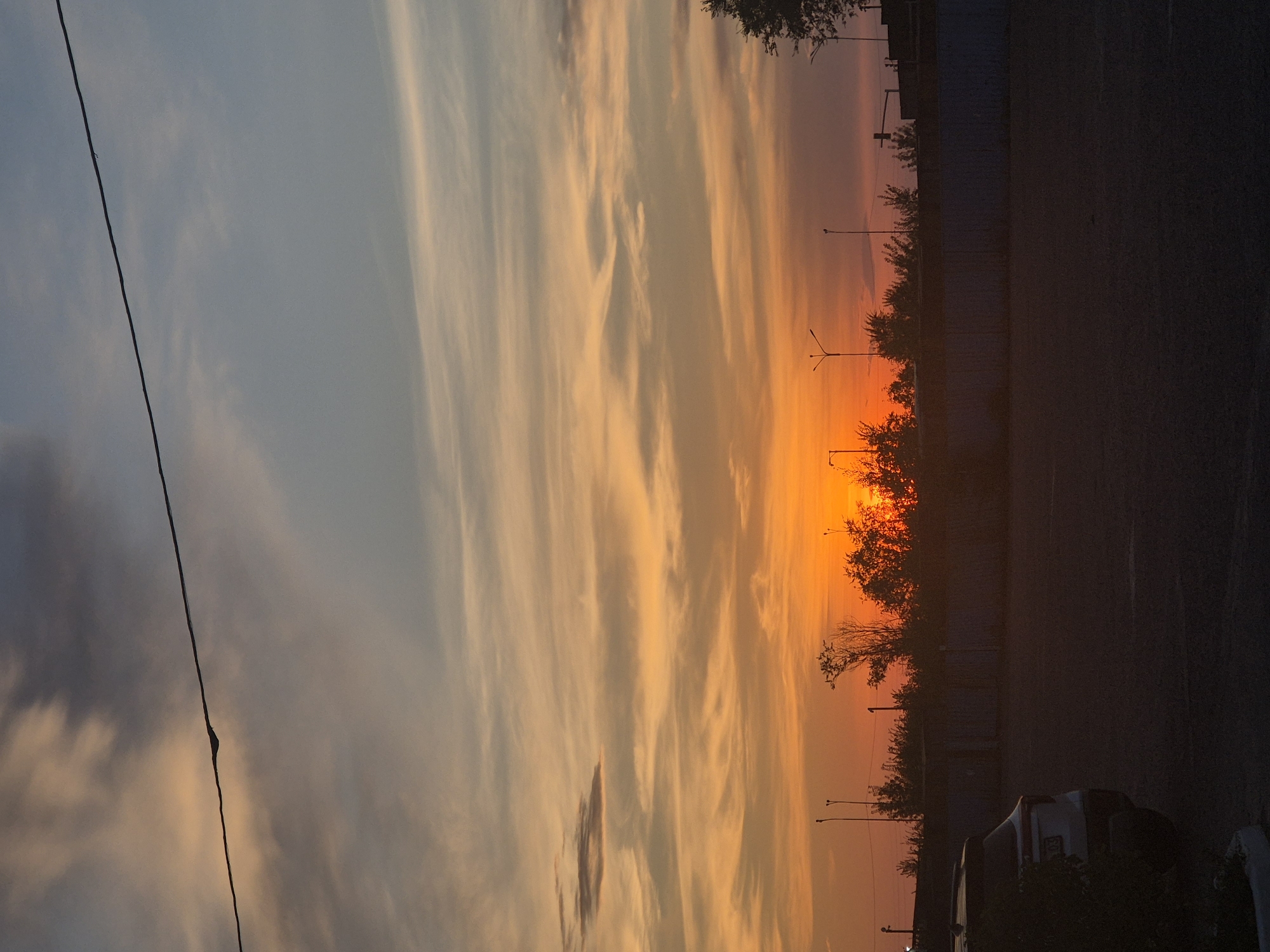 The image captures a sunset scene with a dramatic sky over Kokshetau, Kazakhstan. The sky displays a gradient of colors, with vibrant oranges and yellows near the horizon, transitioning to soft pinks and blues, and finally to a dusky grey at the top, punctuated by wispy clouds and a power line stretching across the frame. Below the sky, silhouetted trees and utility poles form a dark line against the colorful sky. A low concrete wall runs horizontally, separating the foreground from the background. In the foreground, a dark asphalt area, likely a parking lot, is visible. A white SUV is parked on the left side of the frame, partially obscured by shadows and some foliage. The overall atmosphere is peaceful and serene, typical of an urban sunset.