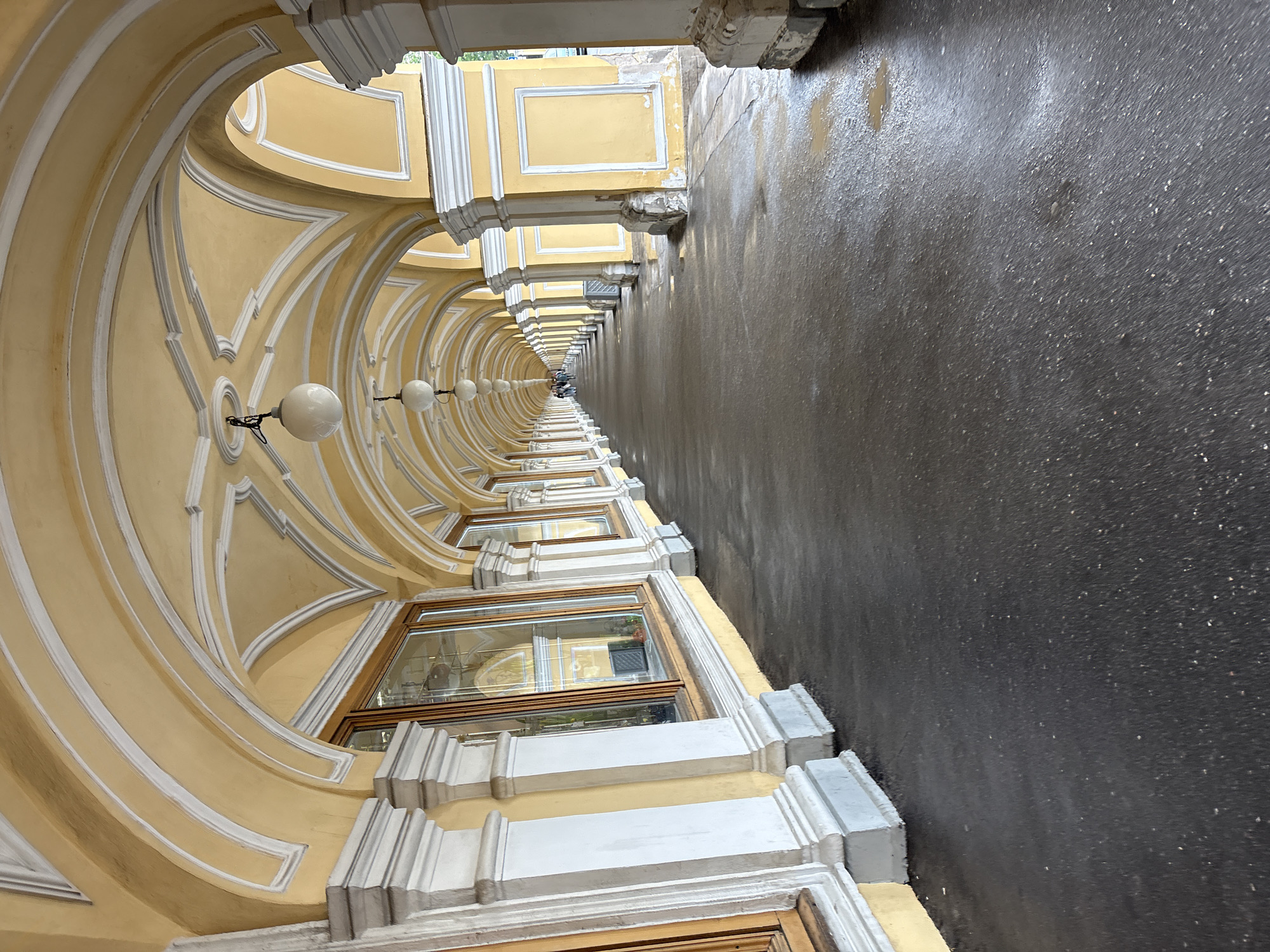 The image captures a perspective view of a covered walkway in Saint Petersburg, Russia. The architectural elements are defined by a series of yellow arches trimmed in white, which extend along the length of the walkway. Spherical white lamps are evenly spaced along the arches, suggesting lighting fixtures. Windows are visible beneath the arches on one side of the walkway. The ground, likely wet asphalt, reflects a dark and grainy texture. At the far end of the walkway, the indistinct figures of one or more people can be seen. The architectural style and location suggest this is part of the State Hermitage Museum complex, specifically along the Winter Canal.