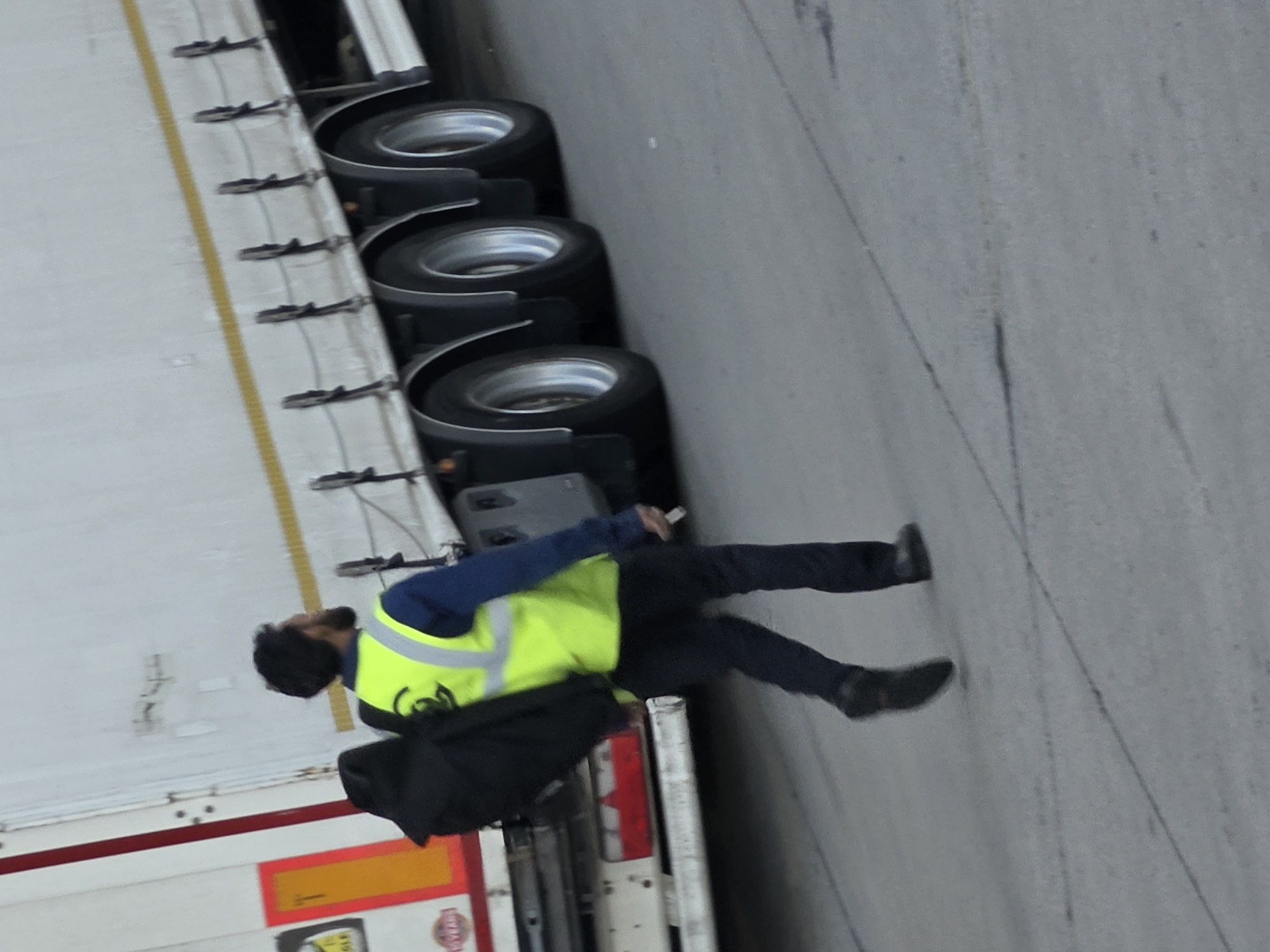 A man in a bright yellow safety vest and dark pants walks away from the camera, carrying a black bag over his left shoulder. He is walking on a grey asphalt surface next to a large white semi-trailer truck with multiple axles. The truck has red and orange reflective markings on its rear. The scene appears to be an industrial or commercial area, possibly a loading dock or depot, in Walldorf, Germany. The lighting suggests it is daytime.