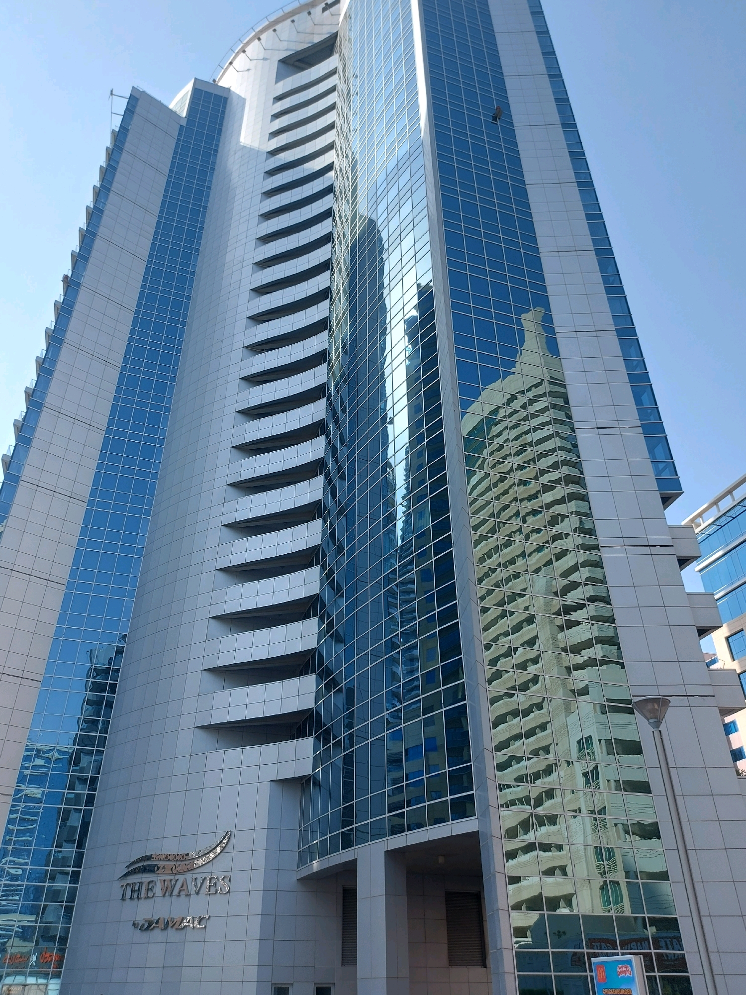 The image shows a low-angle view of The Waves by DAMAC, a residential skyscraper located in Dubai Marina, Dubai, United Arab Emirates. The building features a modern architectural design with a combination of glass and metallic cladding. Several floors have balconies or recessed areas that create a stepped effect on the facade. The glass exterior reflects the sky and neighboring buildings. A street lamp is visible in the lower right corner of the image. A person can be seen on a rope on the exterior of the building, likely performing maintenance or cleaning.
