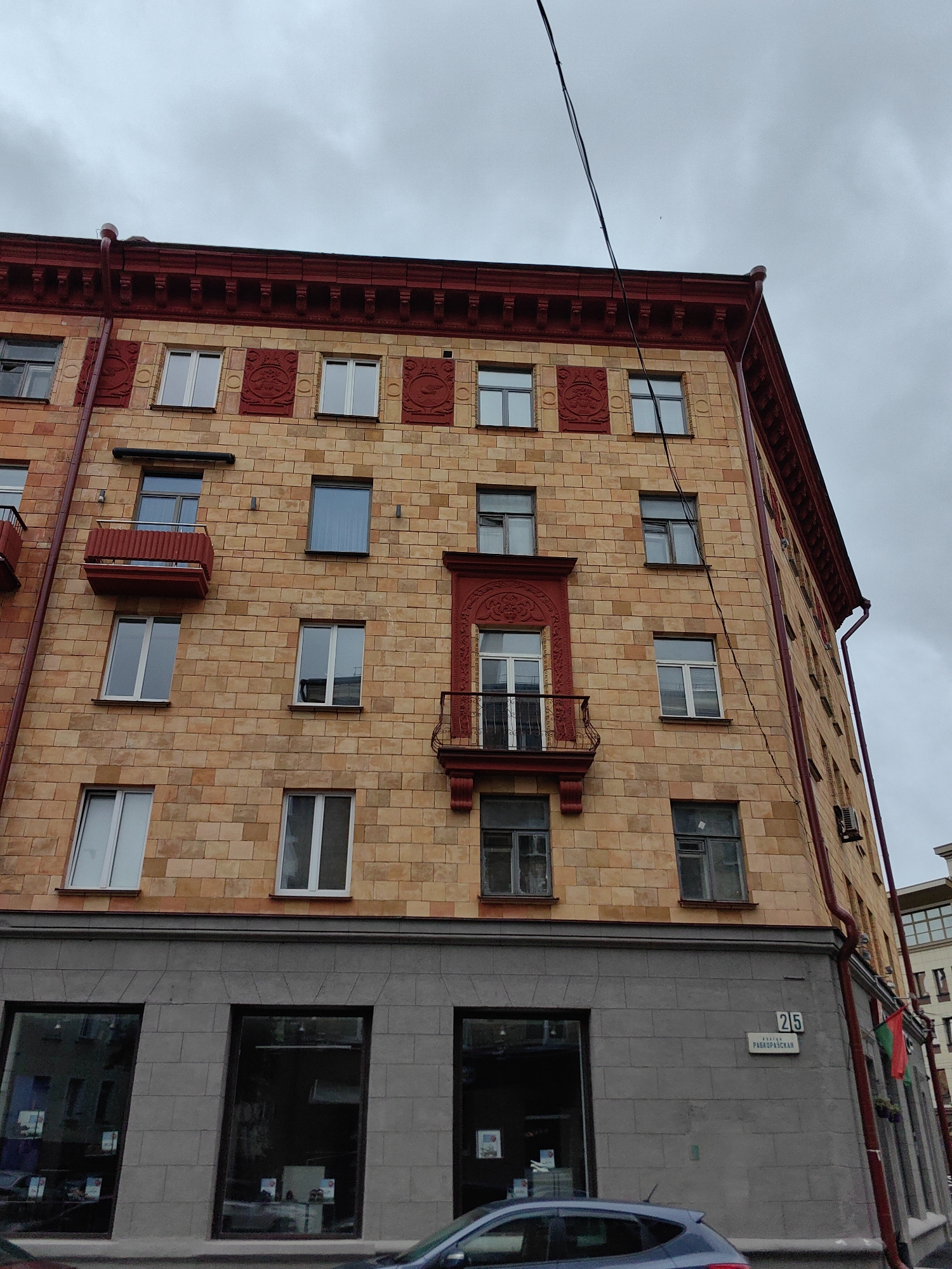 The image captures the facade of an apartment building in Minsk, Belarus, specifically located at **25 Radzivillovskaya Street**. The building has a distinct architectural style, characterized by its beige-toned brickwork interspersed with red decorative elements and red window frames. The upper portion of the building features a red cornice with decorative molding, and several balconies with red railings are visible. The lower part of the building has a gray stone base with large storefront windows. A street sign indicating "25 РАДЗИВИЛЛОВСКАЯ" is mounted on the gray facade. A red downspout runs along the side of the building, and electrical wires are visible against the overcast sky. The scene is devoid of people, but a dark-colored car is partially visible in the foreground.