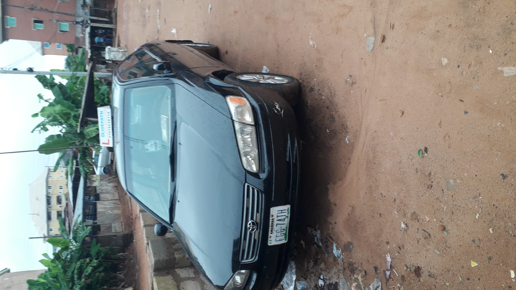 The image shows a black Toyota Camry parked on a dirt road in Nkpor, Nigeria. The front of the car is facing the viewer, and the license plate is visible, reading "ANAMBRA FGG-74TH". The car has a "GLIDEPATH" sign on its roof. The surrounding environment includes banana trees, buildings, and general street debris. The overall impression is of an ordinary street scene in a Nigerian city.