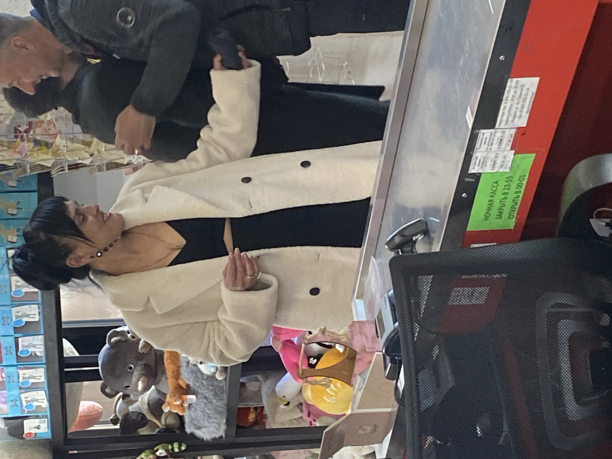 The scene is a busy retail checkout in what appears to be a shop selling toys and other goods, possibly located in Yekaterinburg, Russia, based on the Russian signage. A woman with dark hair pulled back is facing the checkout counter, wearing a black top and a white coat, interacting with a man who is paying, and the checkout assistant, who is partially visible. The background displays shelves stacked with various toys, including plush animals, and retail goods. The checkout counter is equipped with a scanner and Russian signage with store information, including opening and closing times. A metal office chair is visible in the right foreground.