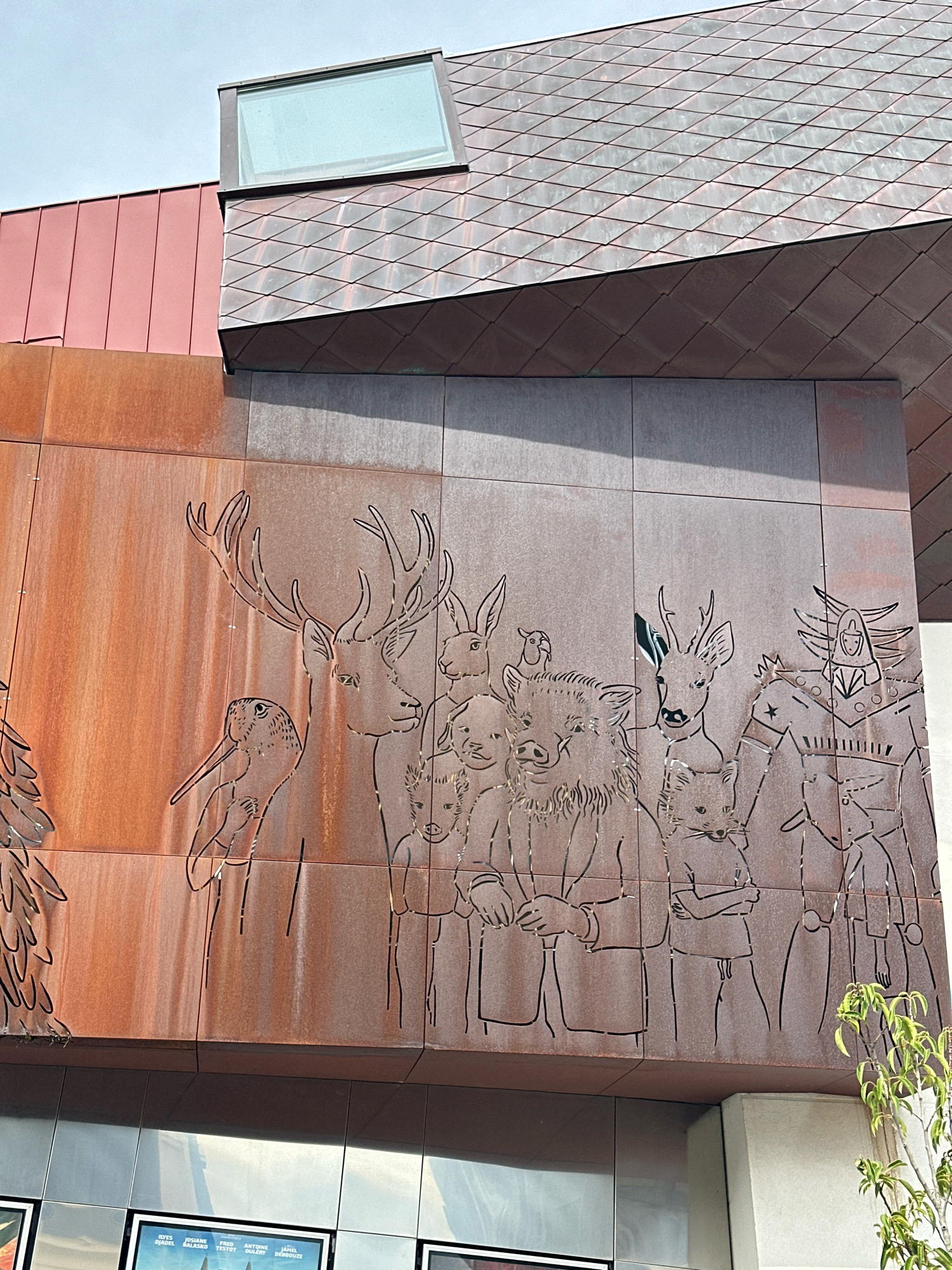 The image shows a close-up of a building facade featuring a metal cutout mural depicting a collection of stylized animals, some of which appear anthropomorphic. The mural is etched into a rusted metal surface. From left to right, the figures include what appears to be a bird with long legs, a large deer with antlers, a rabbit, a pig, a smaller deer, a fox, and a figure with a helmet and armored body, possibly a robot or an armored warrior. Several of the animals are depicted with human-like hands and some are holding objects or have their arms crossed. The building has a modern architectural style with a red vertical panel section and a section covered in diamond-shaped metal tiles. A skylight is visible on the roof. At the bottom of the image, posters or signs are partially visible behind a glass facade. A small green leafy tree branch is visible on the right side of the frame. The scene is set outdoors with a clear sky. The context provided states the location is the city of Roye, France.