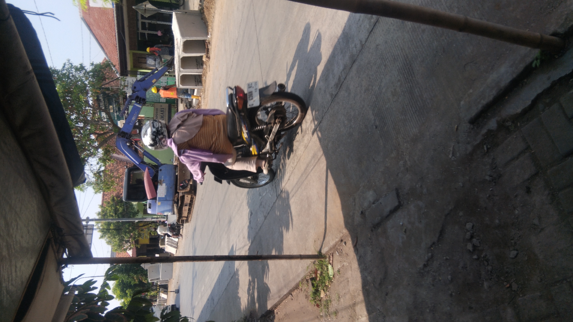 In Batang, Indonesia, on a sunny day, a person wearing a helmet and a purple jacket rides a motorcycle on a paved road. The motorcycle is black with yellow accents. Construction materials, including large concrete pipes, are stacked on the side of the road, next to a blue excavator. The scene appears to be in a residential or commercial area with buildings and trees visible in the background. The road shows signs of wear and tear, with some debris on the pavement.