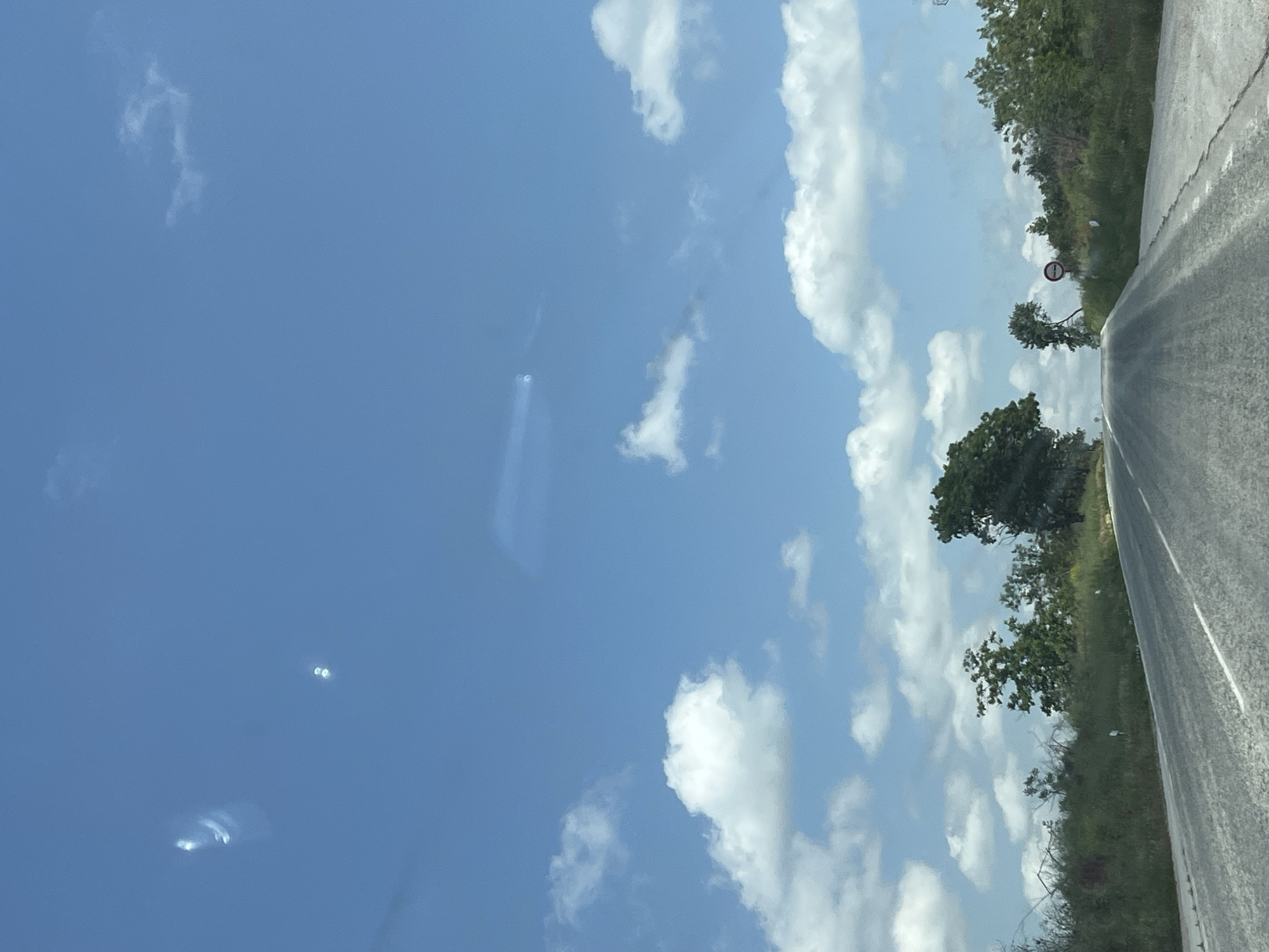 The image shows a view from a vehicle traveling on a road near Kozar Belene, Bulgaria. The road surface is gray and appears slightly wet. The right side of the road is bordered by green vegetation, including trees and bushes. A circular road sign is visible along the roadside. The sky is blue with scattered white clouds. The photo is taken at an angle.