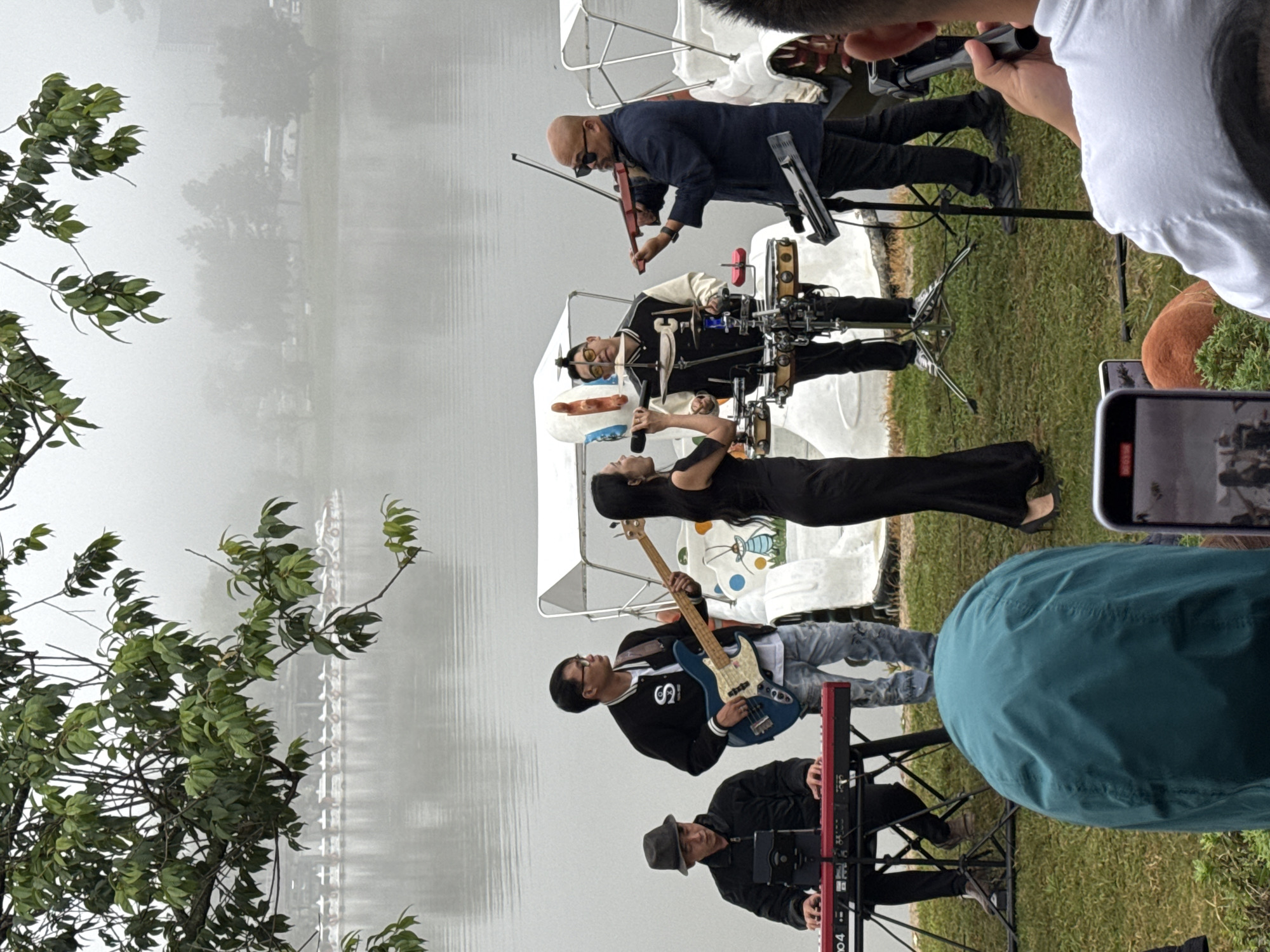 This image depicts an outdoor band performing by a lake in Da Lat, Vietnam. The band consists of four musicians: a keyboardist in a hat on the left, a bassist in glasses in the center-left, a drummer in a letterman jacket in the center-right, and a violinist on the far right. A female singer is also present, holding a microphone in front of the drummer. The band is set up on a grassy area next to the water, with a row of white boats docked in the background. The weather appears overcast, with fog present, obscuring the distant landscape and creating a diffused light. The overall scene captures a live musical performance in a tranquil, natural setting.