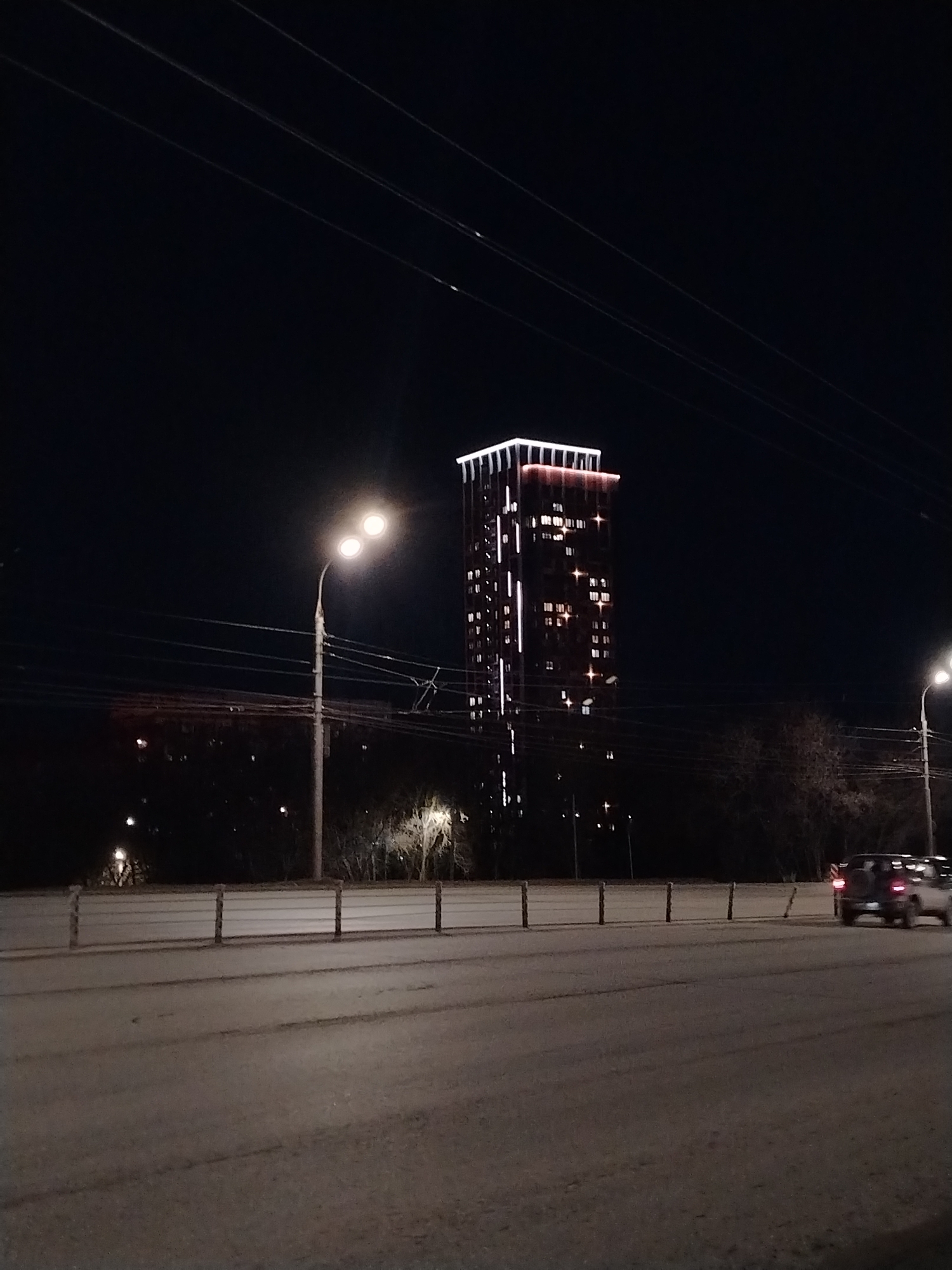 The image shows a nighttime scene in Izhevsk, Russia. A tall, illuminated building stands in the background, with lit windows suggesting it is residential or an office building. In the foreground, a road is visible, possibly a bridge given the presence of a guardrail. A streetlight illuminates the road. A vehicle with illuminated taillights is moving on the road. The sky is dark, and utility lines are strung above the scene. There are some trees and foliage visible in the mid-ground between the road and the building.
