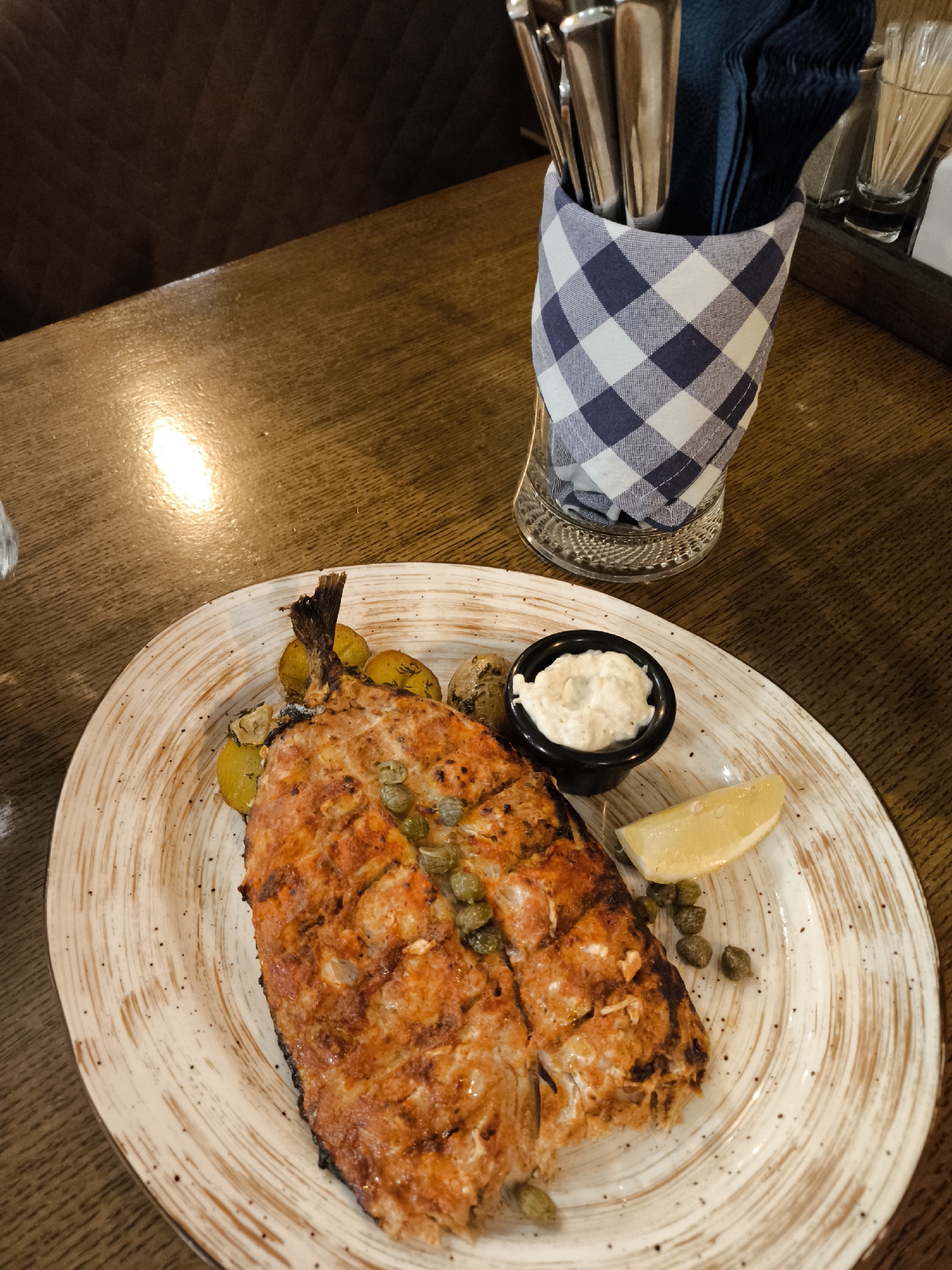 This media file features a close-up, top-down view of a plate of food on a wooden table. The primary subject is a grilled fish, appearing to be salmon, with visible grill marks and a reddish-orange hue. It is topped with capers and served alongside roasted potatoes, a wedge of lemon, and a small dish of white sauce, likely aioli or tartar sauce. The plate itself is a textured, light-colored ceramic with a rustic design.
In the background, a glass container holding cutlery, wrapped in a blue and white checkered cloth, is visible. There are also toothpicks in a holder and a dark blue napkin. The setting appears to be a restaurant, with the warm tones of the wooden table and the overall lighting suggesting an indoor environment. The lighting creates a slight glare on the table surface.
The image focuses solely on the food and its immediate presentation, with no people visible. The activity taking place is implied to be a meal being served or about to be eaten. There are no discernible text elements within the image. The context provided indicates the location is Moscow, Russia, which doesn't offer specific visual cues in this particular crop. The time of day and weather are not determinable from the image alone. The overall impression is one of a well-prepared, appetizing dish in a casual dining setting.