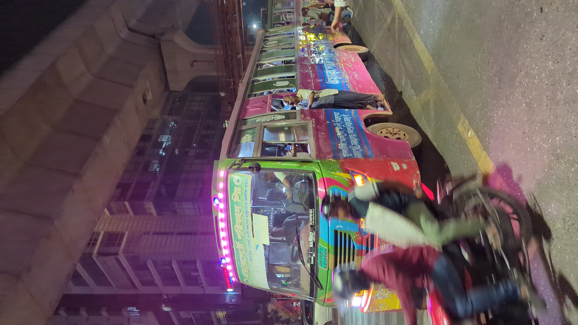 The image captures a vibrant scene in Dhaka, Bangladesh, featuring a colorfully decorated bus at night. The bus has a green, red, and pink exterior and is adorned with illuminated LED lights on the roof. People are visible on and around the bus, with one individual resting on the bus's side. Some other people are riding motorbikes passing the bus. The setting appears urban, with a large elevated highway structure and high-rise buildings in the background, suggesting the location is within Dhaka. The street is illuminated with lights, and shadows indicate it is nighttime.