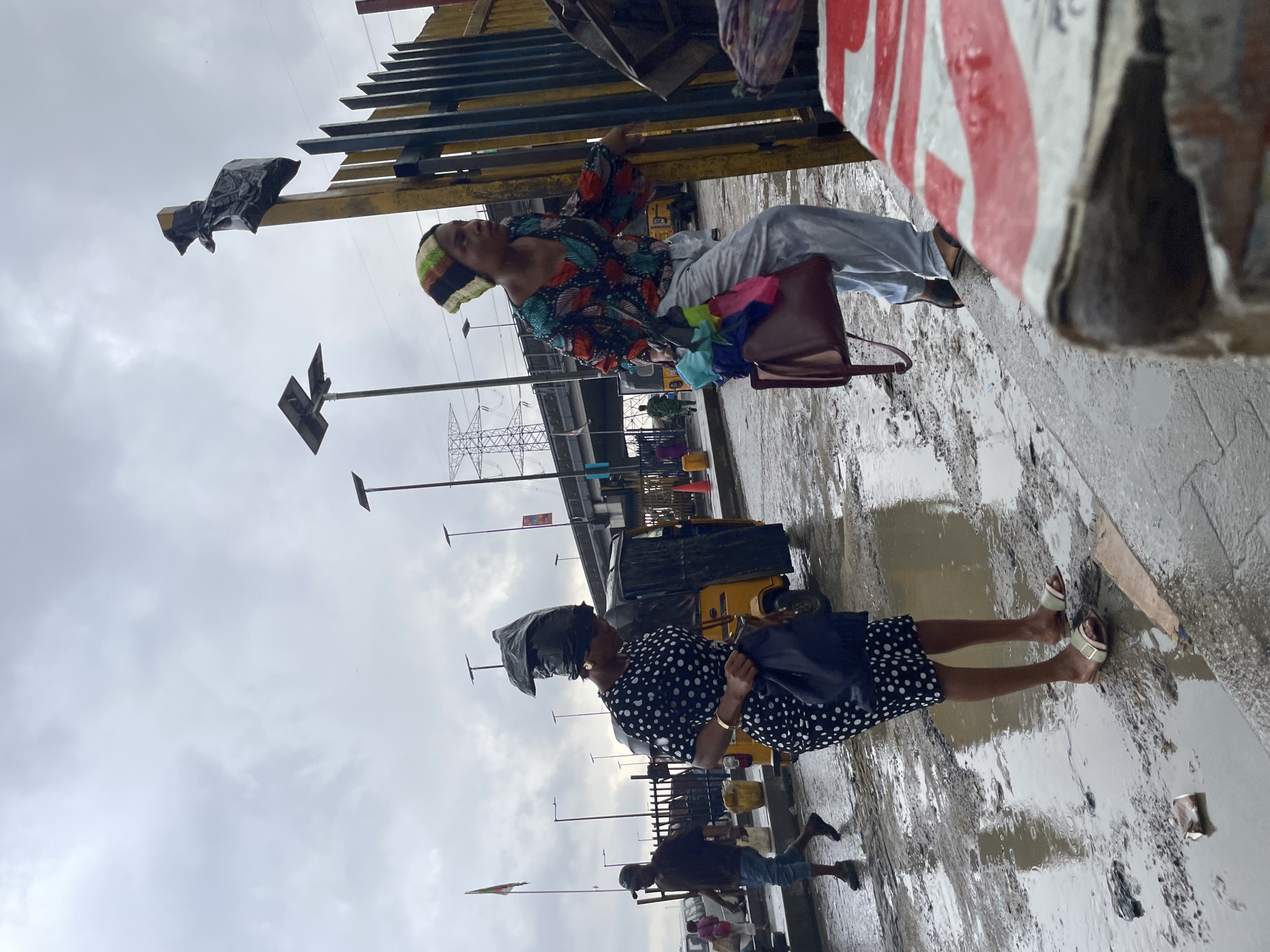The image depicts a street scene in Ado, Nigeria, under a cloudy sky. In the foreground, a woman in a black and white polka dot dress walks through puddles on a wet, uneven road, carrying a dark bag and wearing a black plastic bag as a head covering. To her right, another woman in a colorful patterned shirt and light-colored pants walks, holding a burgundy handbag and a bundle of colorful fabrics. Behind them, a yellow auto-rickshaw is parked. In the background, more people are visible, along with streetlights, a flag on a pole, and electrical infrastructure including power lines and a transmission tower. The road is heavily puddled, indicating recent rainfall or a poorly drained area. A fence, painted yellow and black, is prominent on the right side of the image. The overall impression is one of a busy, potentially impoverished urban environment during or after a period of rain.