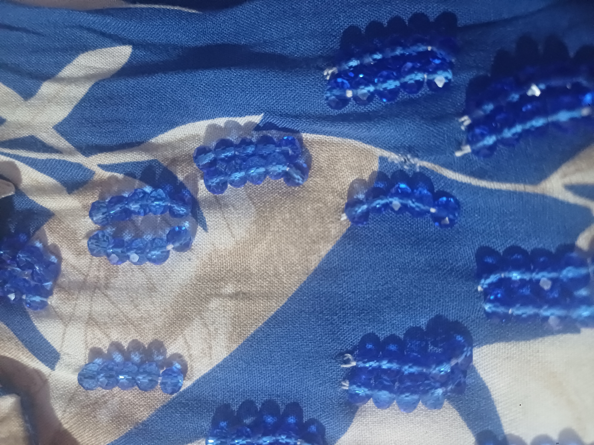 The image shows a close-up view of blue beads arranged in clusters on a patterned fabric. The fabric has a background of beige and white with blue stripes and abstract shapes. The beads are faceted and appear to be made of glass or plastic, reflecting light. Some of the bead clusters are elongated, while others are more compact. The image is taken from a high angle, and the lighting creates shadows that accentuate the texture of the beads and fabric. There is no information in the image to suggest a location in Jumashuy, Uzbekistan.
