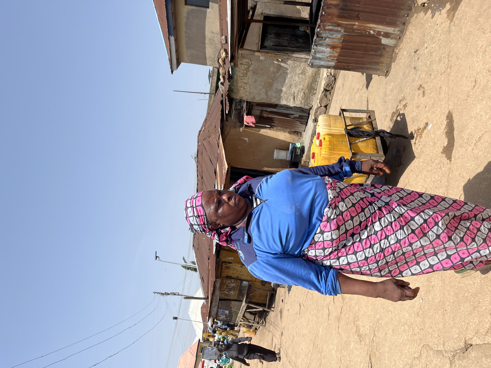 A woman is captured in a medium shot, standing on a dusty street in a market setting. She is wearing a bright blue long-sleeved shirt and a colorful wrap skirt with a pink and white geometric pattern. Her head is covered with a pink and white patterned headscarf. Her gaze is directed slightly to her left, and her expression appears calm.
Behind her, several large yellow plastic containers are stacked on a metal trolley, suggesting she might be a vendor or involved in transporting goods. The background reveals a bustling marketplace with simple, weathered structures, some with corrugated metal roofs. These buildings appear to be shops or stalls. There are other people visible in the distance, as well as some market activity. The ground is unpaved and appears dry and dusty.
The scene is bathed in bright sunlight, indicating a daytime setting, likely a warm day. The clear blue sky suggests fair weather. The overall impression is one of everyday life in a Nigerian market. A partial logo on the woman's shirt is visible, featuring the word "DOMINO'S".