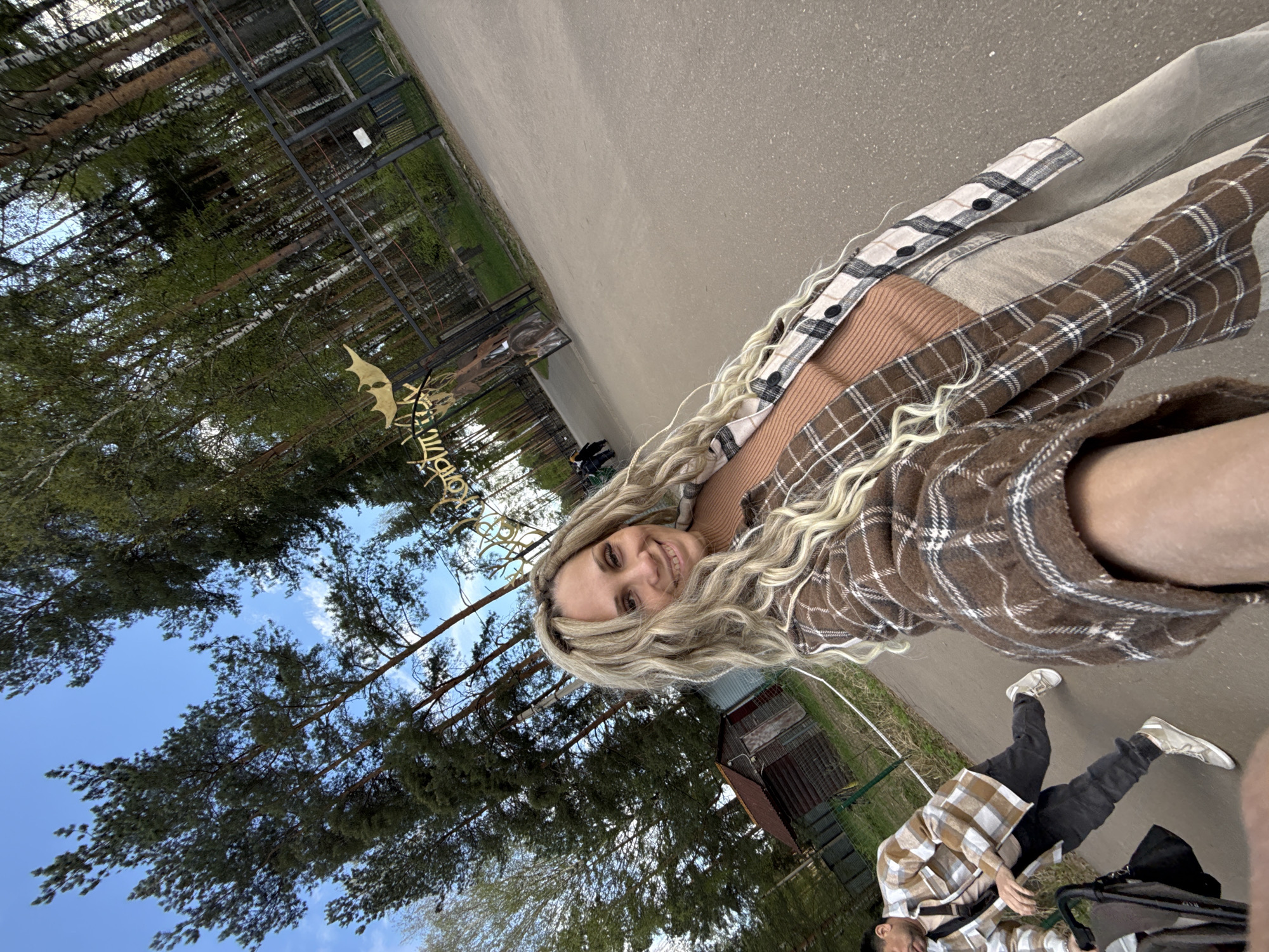 A woman with blonde dreadlocks smiles in a selfie taken outdoors in a park. She wears a tan top and a layered plaid shirt. Behind her, a path leads away, lined by tall trees and greenery. A man is visible pushing a stroller, wearing a plaid shirt and dark pants. A decorative sign is partially visible in the background, partially obscured by trees, indicating the location is likely in Yaroslavl', Russia. The sky is blue with some clouds.
