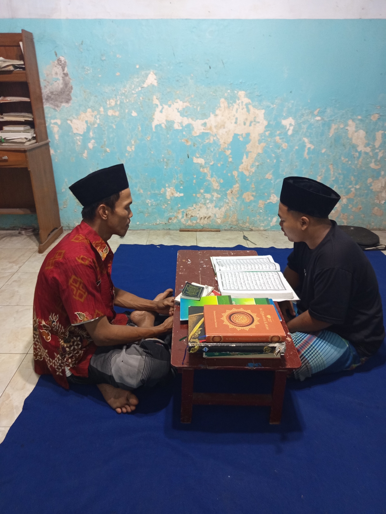 In a dimly lit room with a peeling blue and white painted wall, two men wearing black peci hats are seated on a blue mat. They are positioned opposite each other, facing a small, dark red wooden table. The man on the left, wearing a red shirt with gold patterns, is looking towards the man on the right. The man on the right, dressed in a black t-shirt, is looking at an open book on the table. Several books are stacked on the table, including what appears to be a Quran. A wooden bookshelf with stacks of papers is visible on the left side of the image. The scene suggests a religious or educational setting, likely a private residence or a small religious institution in Ngablak, Indonesia.