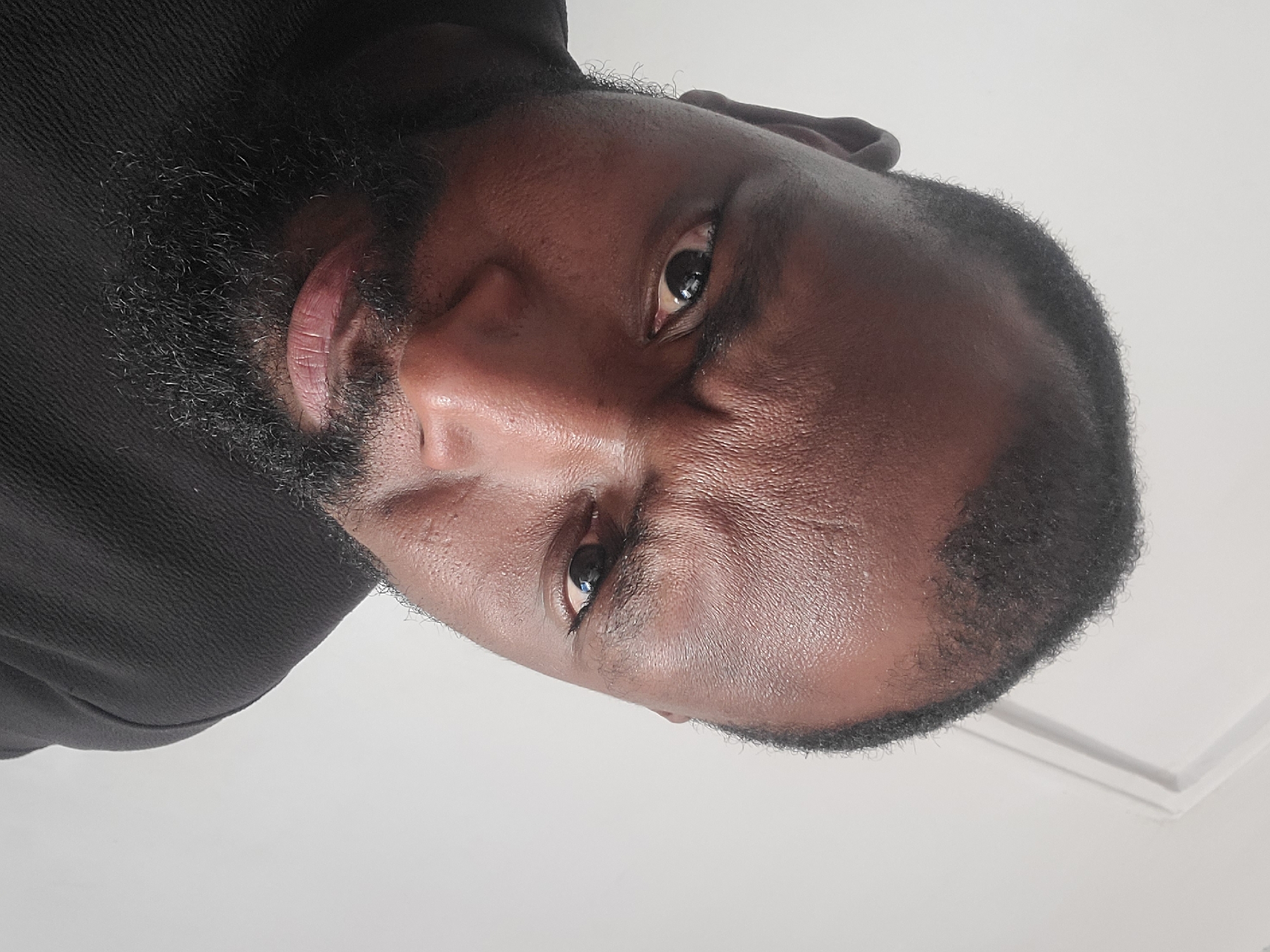A close-up portrait shows a man with dark skin, a full dark beard, and short dark hair, looking directly into the camera. The image is oriented sideways, suggesting the person is lying down and looking upwards, or the image has been rotated from its original vertical or horizontal capture. He wears a dark, possibly black, textured garment, visible around his shoulder and neck. His eyes are dark brown, and his gaze is direct and neutral. The lighting is bright and even, highlighting the texture of his skin and beard. The background is a plain, light-colored surface, likely a white wall or ceiling, with a subtle architectural edge visible in one corner, indicating an indoor setting. There is no visible text. The image is likely taken in Jos, Nigeria.