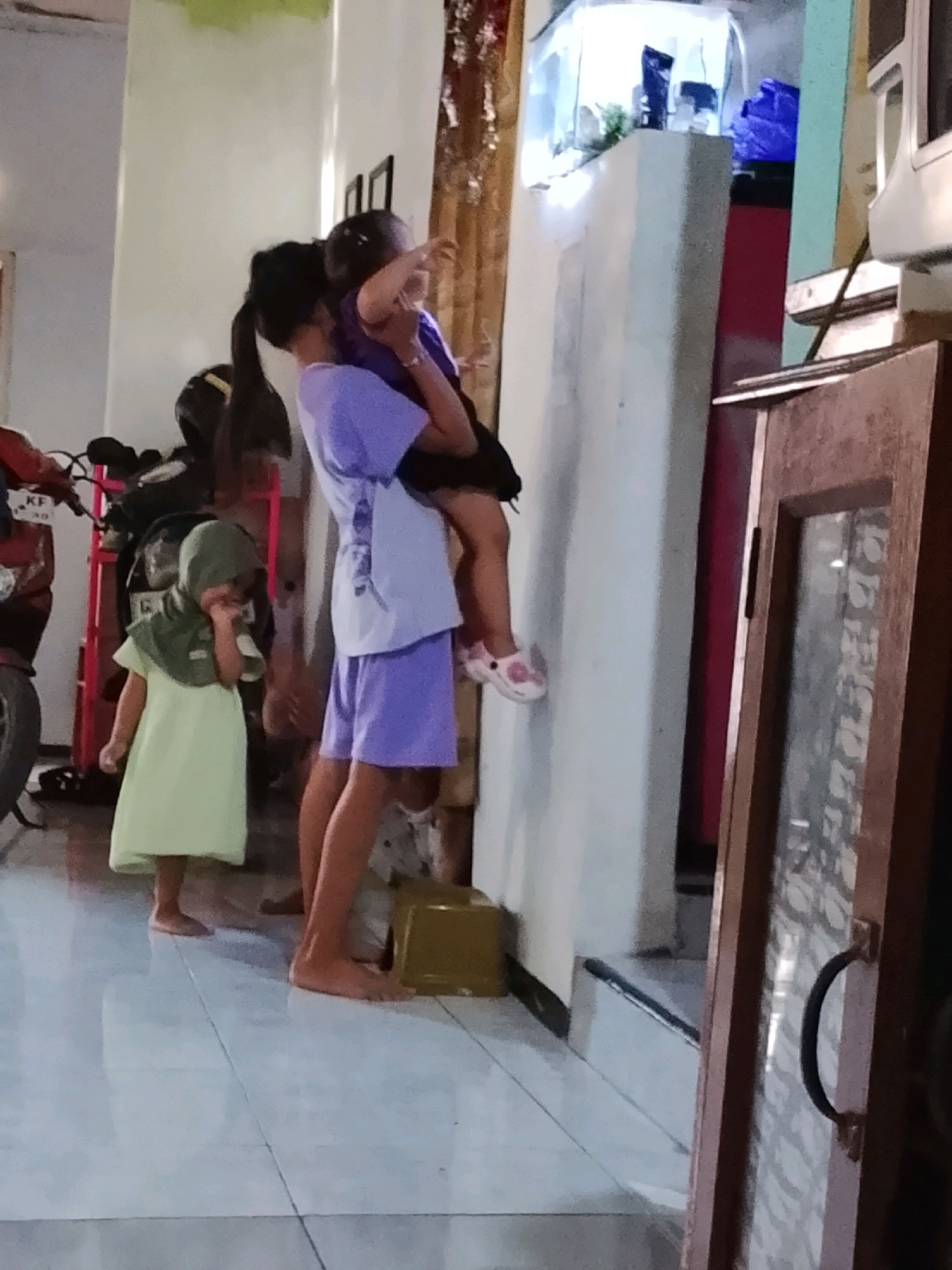 In an indoor scene, likely a home in Ujungrusi, Indonesia, a young girl in a light green dress and green hijab is standing on a tiled floor, reaching for something with her hand. She appears to be looking down at her hand or something near it. To her right, an older girl in a purple t-shirt and purple shorts is holding a baby. The baby, facing away from the viewer, is wearing a dark purple top. The older girl is holding the baby at chest level, with her arms supporting the baby's back and legs. The older girl's legs are visible, with one foot bare and the other wearing a pink shoe with a floral pattern. The background reveals a motorcycle, a red shelf, and a tall, light-colored structure, possibly a cabinet or aquarium stand, with a blue object on top. Further to the right, a portion of a wooden cabinet with a glass door and a dark handle is visible. The lighting suggests an indoor environment, possibly during the day.