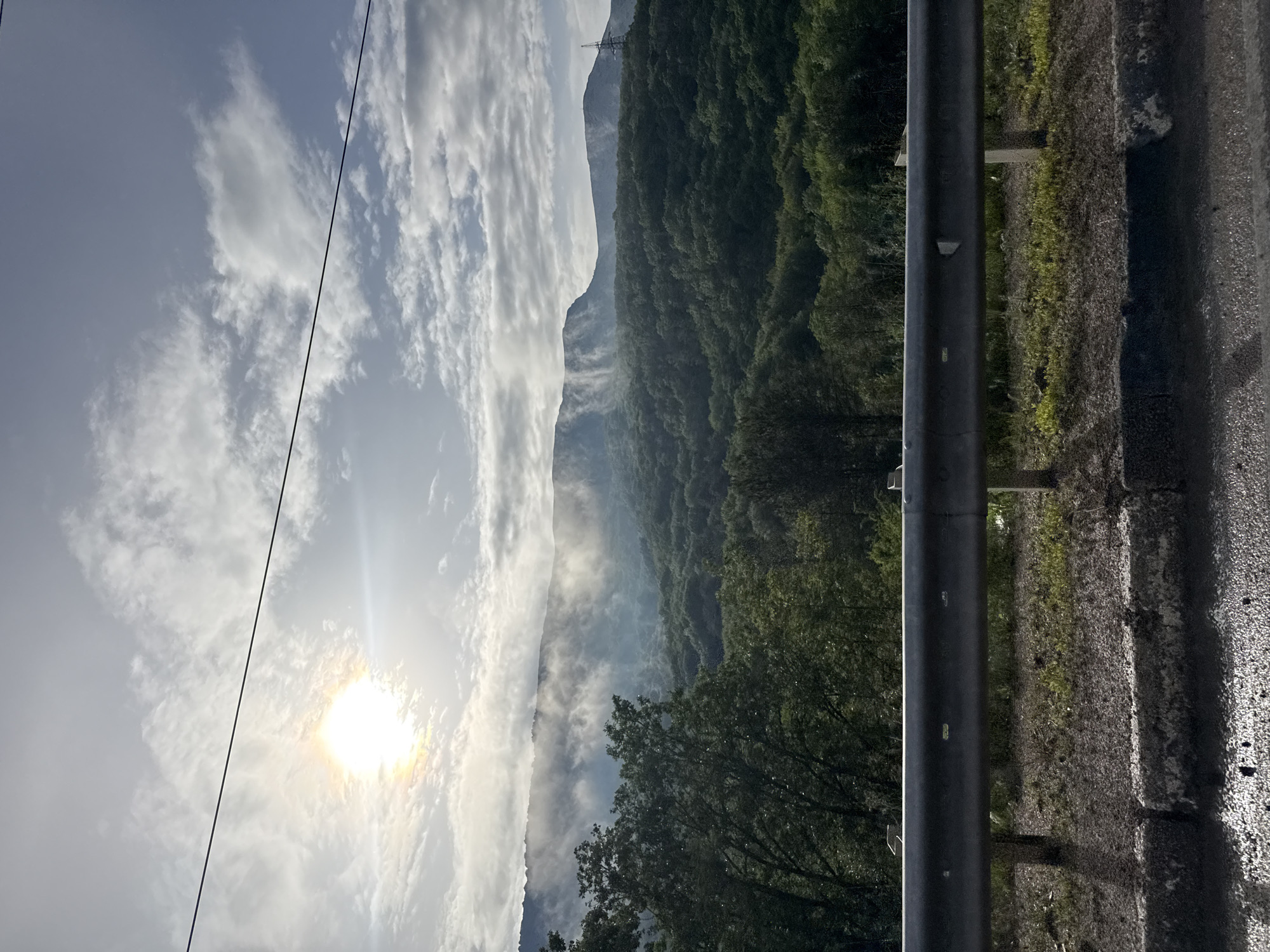 A horizontal, eye-level shot captures a serene mountain landscape under a partially cloudy sky. The sun, a bright, diffused orb, peeks through the clouds on the left side of the frame, casting a soft glow and creating lens flare. A thin, dark power line stretches diagonally across the upper portion of the image.
In the mid-ground, rolling hills are covered in dense, lush green trees, their canopies forming a textured carpet. Wisps of fog or mist gently embrace the upper slopes of the mountains, adding a layer of mystery and depth. The mountains themselves are vast and rugged, their peaks silhouetted against the sky. On the far right of the mountain range, a tall, lattice-like power transmission tower is visible.
In the foreground, a grey metal guardrail runs horizontally, acting as a barrier between the road and the hillside. It is supported by posts that are partially obscured by vegetation. The ground beneath the guardrail consists of dirt and sparse, dry grass. The very bottom of the image shows a wet, asphalt-like surface, possibly a road, with reflective patches suggesting recent rain. The overall scene evokes a sense of natural beauty and tranquility, perhaps captured from a roadside vantage point.