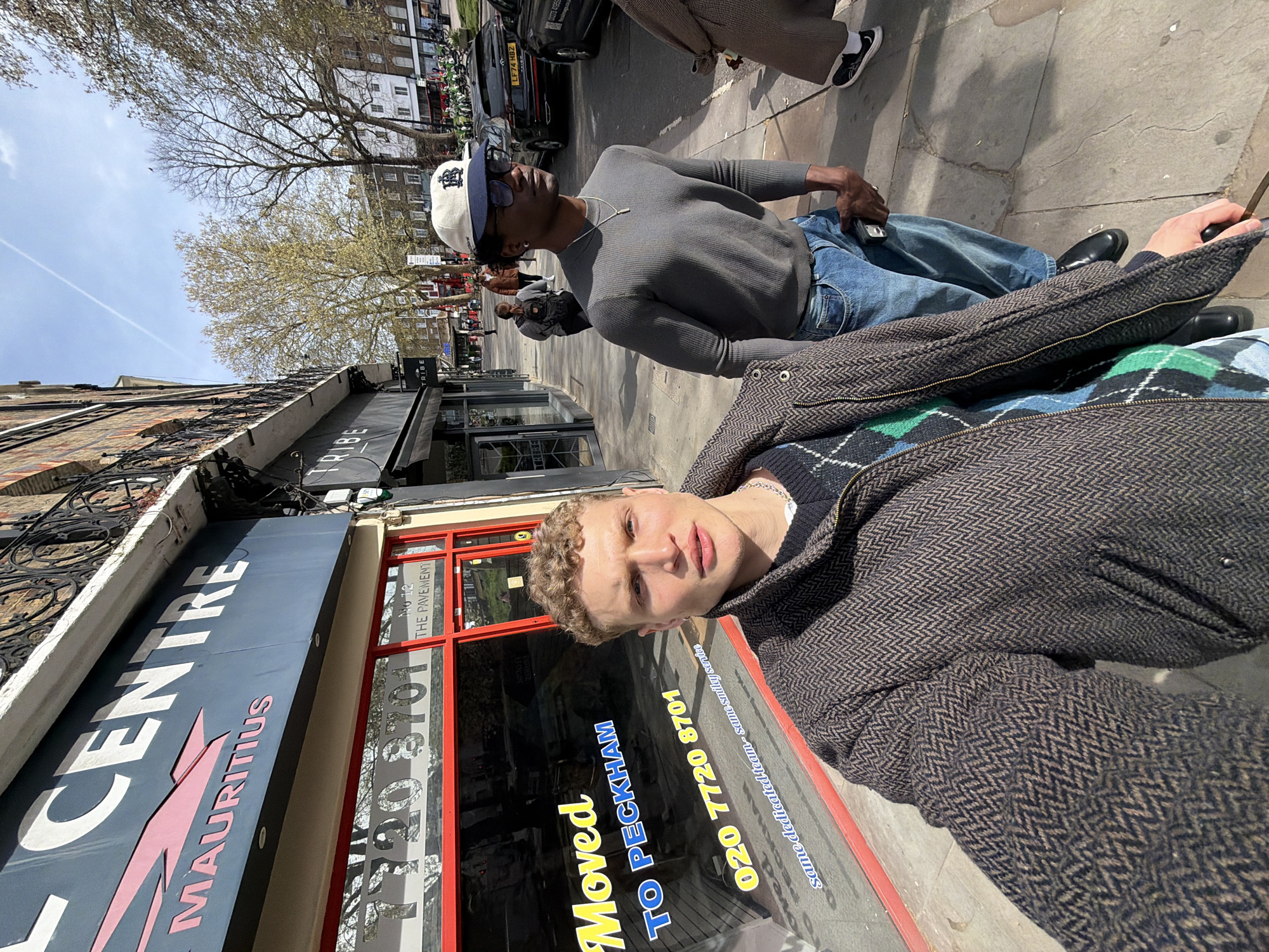Two men stand outside of a building on a street in London, United Kingdom. The blonde man in the foreground faces the camera, wearing a textured brown jacket and a blue and green patterned sweater. The second man wears a gray shirt, jeans, black shoes, and a white baseball cap. Behind them, cars are parked along the street. Buildings are visible, including one with a sign for "CENTRE MAURITIUS." A sign in the window reads "Moved TO PECKHAM 020 7720 8701." A store called TRIBE is located across the street.