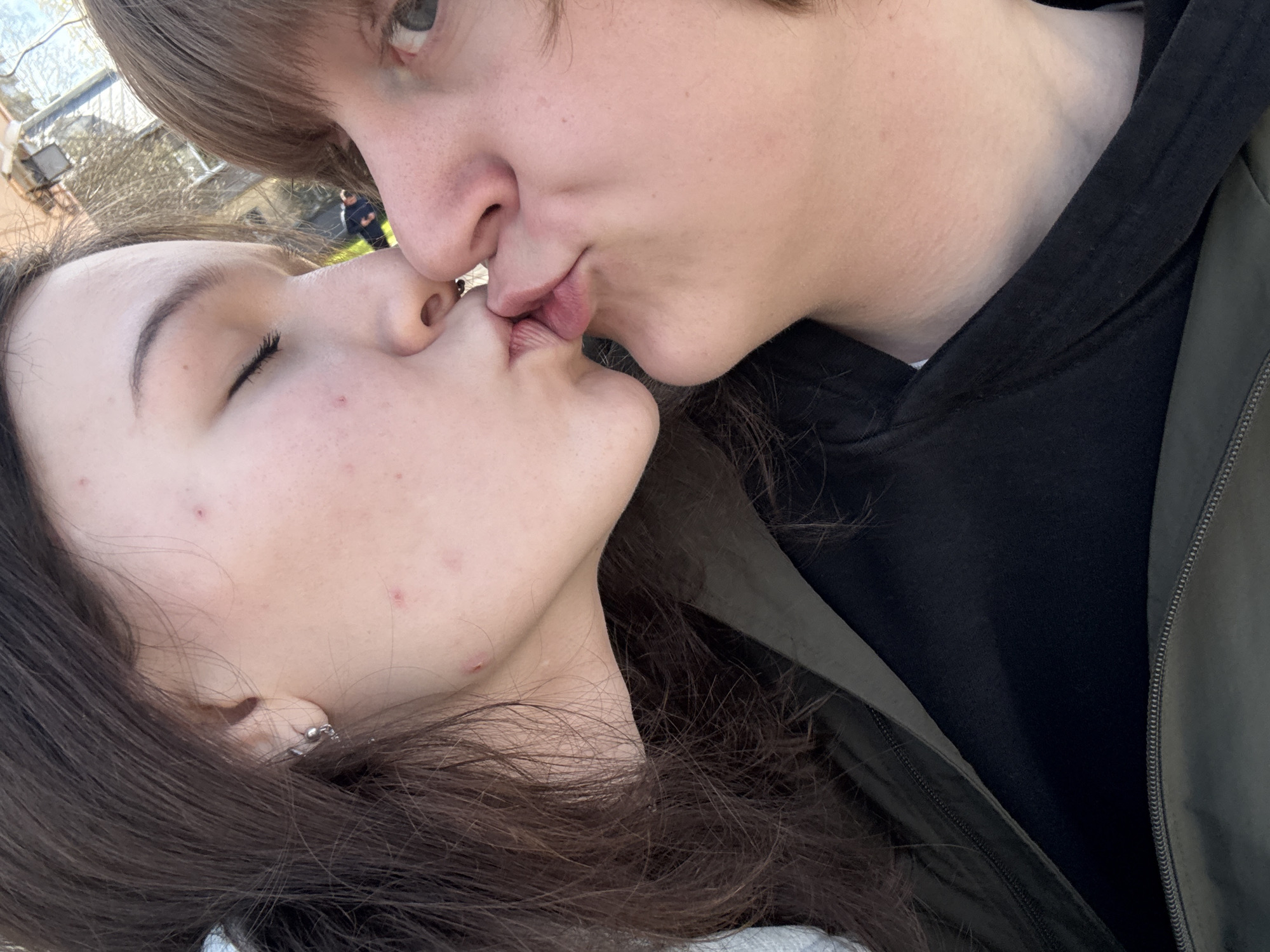 An intimate close-up shot captures two young people kissing outdoors in Vsevolozhsk, Russia. The image is taken from a slightly tilted angle, likely a selfie, emphasizing their affectionate interaction.
The female subject is positioned in the foreground, with her profile clearly visible. She has long, dark brown hair that frames her face and flows over her shoulder. Her eyes are closed, indicating a moment of tenderness during the kiss. Several small red acne spots are visible on her cheek, forehead, and jawline. She wears a small earring in her ear and a dark green jacket layered over a lighter garment.
The male subject is partially visible on the right side of the frame, positioned above the female. His upper face, including his nose, mouth, and one light-colored eye, is prominent. His light brown hair partially covers his forehead. He wears a dark, possibly black, hoodie underneath what appears to be an olive green or dark jacket, with a visible zipper seam on the right.
Their lips are pressed together in a tender kiss. The background is softly blurred, showing hints of outdoor elements such as trees or distant buildings under natural daylight, suggesting a clear day. There is no visible text in the image.