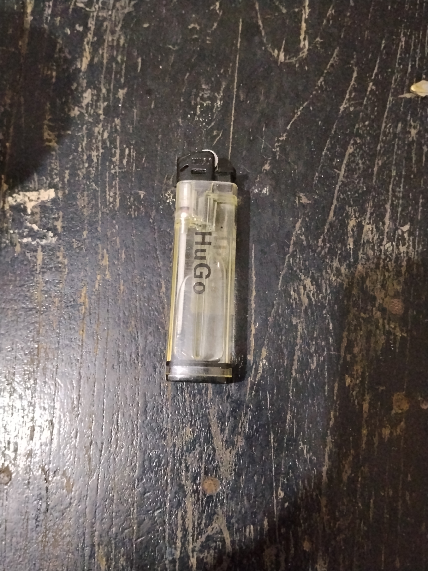 A clear plastic lighter with the brand name "HUGO" printed vertically on its side lies on a weathered, dark-colored wooden surface. The wood shows signs of age and wear, with visible grain and paint peeling in places. The lighter is positioned slightly off-center to the right. The lighting appears to be dim, casting subtle shadows. No people or actions are depicted. The environment suggests an indoor or outdoor surface that is likely exposed to the elements or heavy use. There is no information about the location in Bilis Bilis, Indonesia.