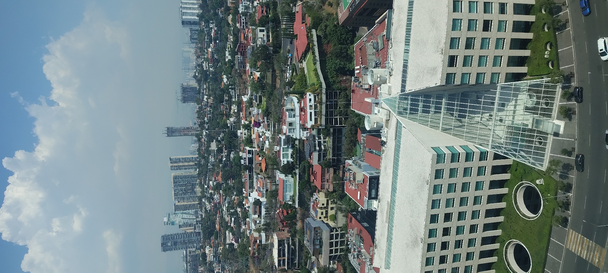 This image shows a daytime aerial view of a portion of Mexico City, Mexico. The urban landscape is characterized by a mix of residential and commercial structures.
To the left of the image, numerous buildings of varying heights, likely apartments and commercial buildings, stretch towards the horizon, partially obscured by a hazy sky. A large cloud formation fills the upper left corner of the image.
In the central section of the image, dense residential areas are visible, featuring houses of diverse architectural styles and colors, interspersed with green spaces and trees.
The right side of the image is dominated by a tall, modern building with a glass facade, potentially an office or residential tower. Below the building, there's a well-maintained green area with circular designs and a street with cars parked along the side.
Overall, the image captures the diverse urban fabric of Mexico City, showing a blend of residential neighborhoods and modern high-rise buildings.