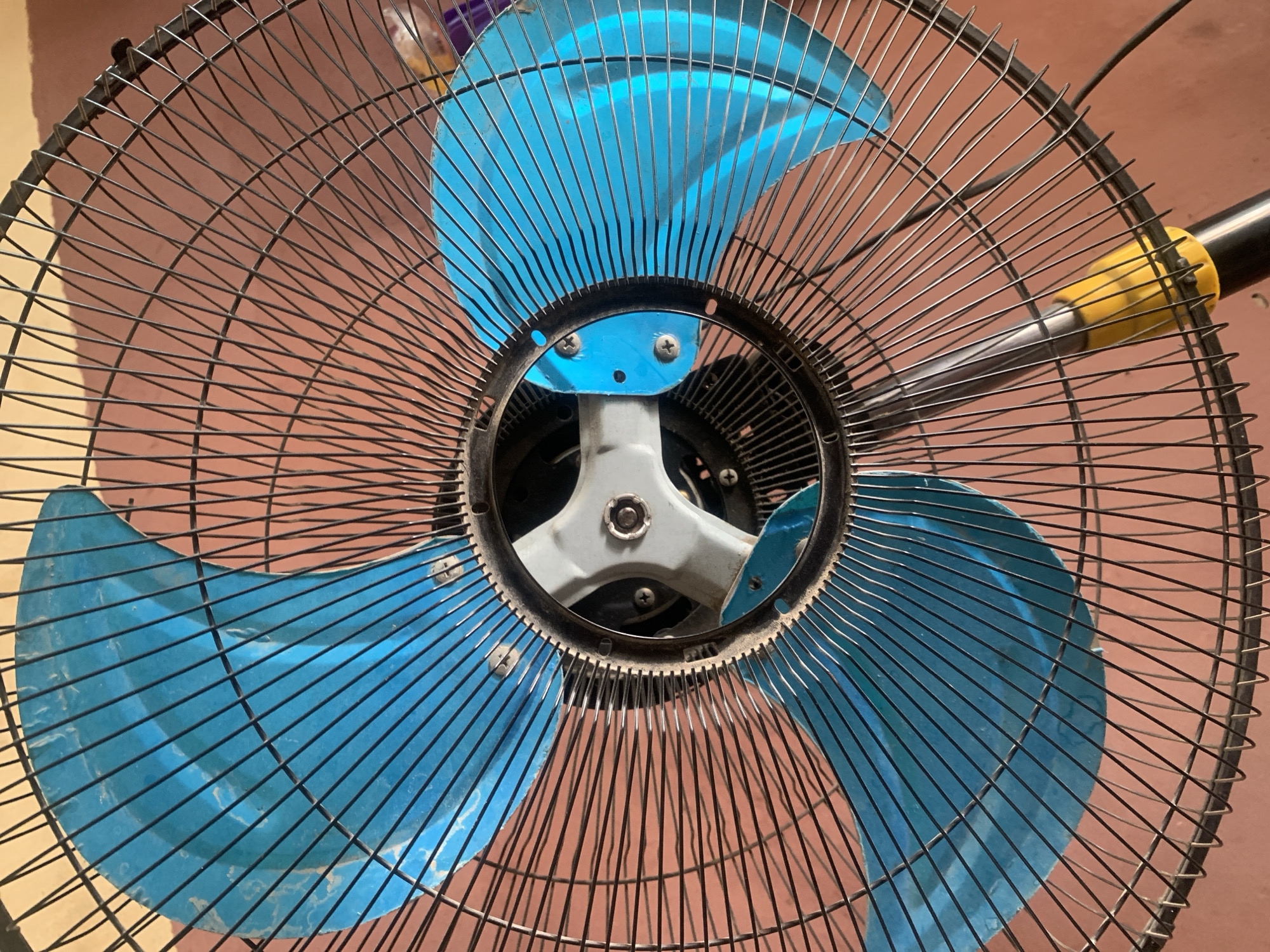 A close-up, low-angle shot shows the front grille and blue blades of a standing fan. The fan's wire mesh grille, made of thin black wires, covers the entire frame. The three blue fan blades are visible through the grille. The center of the fan has a metallic hub with a plastic component. The background is a reddish-brown wall. The fan is positioned indoors. The location in Jauro Yinu, Nigeria is unknown.