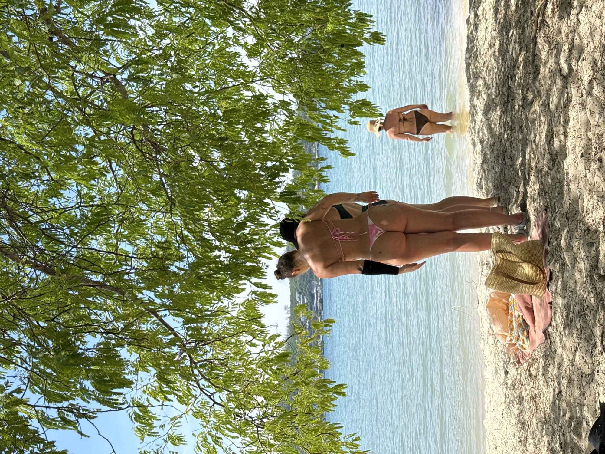 In Nha Trang, Vietnam, on a sandy beach partially shaded by a tree with green foliage, three women are present. Two women, facing away from the camera, stand near the waterline. The woman in the foreground on the left wears a pink bikini. Beside her, another woman also wears a bikini. In the background, a third woman wearing a dark bikini and a straw hat stands in the shallow ocean water. On the sand in the foreground, a beach bag and folded towels are visible. The ocean is calm and a hazy horizon with buildings and vegetation is discernible. The sky is clear blue.