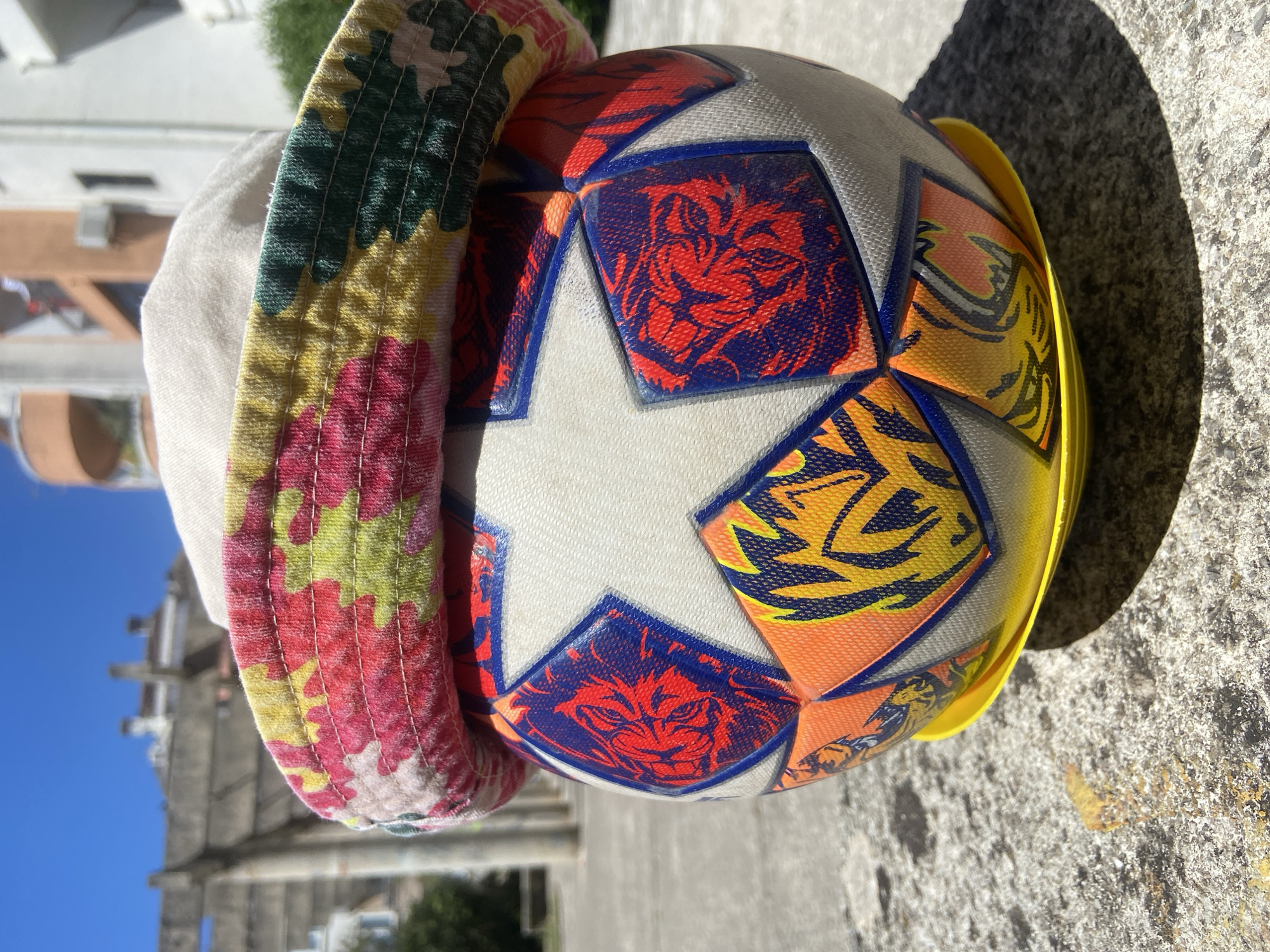 A close-up shot depicts a soccer ball placed on a concrete surface outdoors. The ball is partially covered by a bucket hat with a floral pattern on its brim and a plain white crown. The soccer ball features a design of stylized tigers in red and blue on a white background with a yellow plastic rim. The background features a building facade and a clear blue sky, indicative of an outdoor location. Based on the architecture and sky, this scene appears to be set outdoors in an urban area near historic buildings, potentially located in the city of Budva, Montenegro.