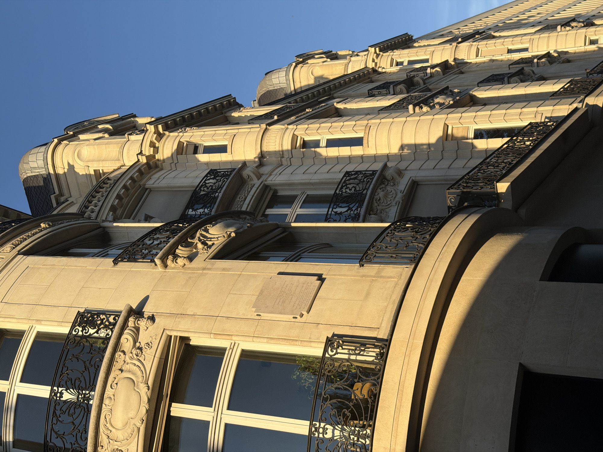 The image showcases the architecture of a building in Paris, France, likely a residential or commercial structure. The building features a classic Parisian style with detailed stone facade, ornate ironwork balconies, and multi-pane windows. The low-angle perspective emphasizes the building's height and the ornate design elements. The clear blue sky in the background suggests a sunny day.