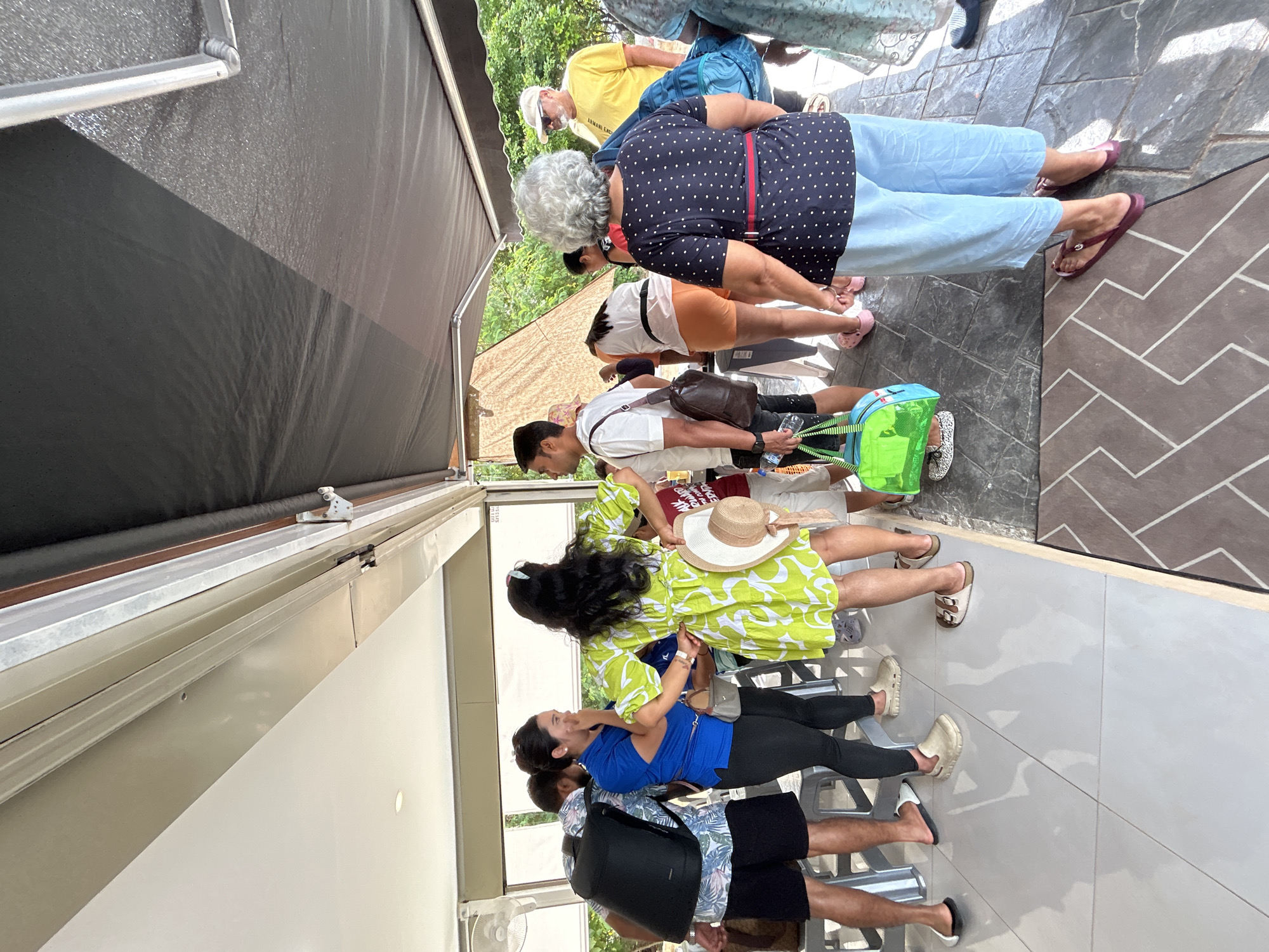 A group of people are gathered outdoors near a building, possibly a restaurant or cafe, in Ratsada, Thailand. The scene is partially covered by a dark awning. In the foreground, a woman in a lime green dress with white floral patterns is holding a straw hat. Next to her, a woman in a blue shirt and black leggings is speaking, with her hand near her mouth. A man wearing a black backpack and a floral shirt stands to their left. Further back, several other individuals are visible, some holding bags and others standing and observing. The ground is tiled with a pattern of light and dark grey squares. Trees and vegetation are visible in the background, suggesting a tropical environment. The overall impression is a casual gathering of people outdoors.
