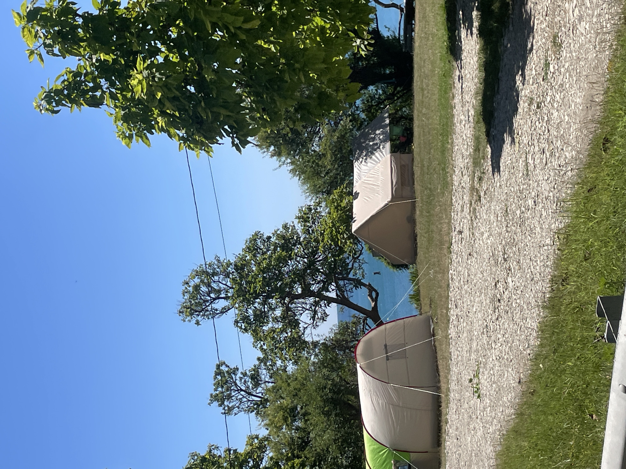 The image depicts a campground setting with two tents pitched on a grassy area. The foreground is a path covered in small stones, with patches of green grass on either side. Beyond the tents, a body of water, likely the sea, is visible under a clear blue sky. Several trees with green foliage are scattered throughout the scene, with a prominent large tree on the right side of the frame. No people are visible in the image. The location is identified as a campground in Dzhubga, Russia.