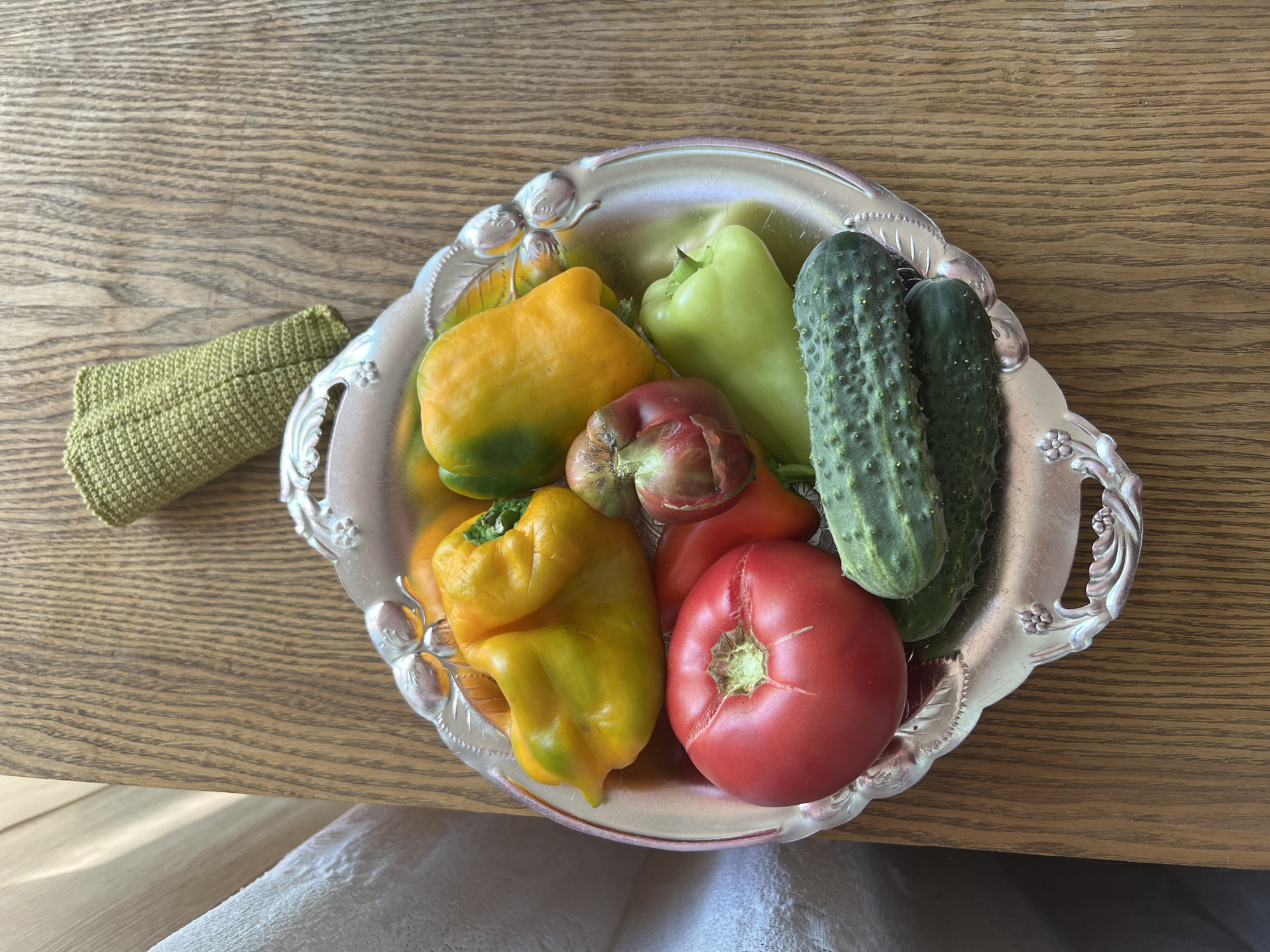 A still life arrangement of fresh produce is displayed on a decorative silver platter. The platter contains a large, ripe red tomato, two dark green cucumbers, and several bell peppers in various stages of ripeness, ranging from bright yellow with green accents to solid green. A small, deeply colored heirloom tomato with ribbed skin is also visible. The platter sits on a wooden surface, likely a table, with a textured green crocheted item positioned to the upper right. A translucent white fabric is draped to the left of the platter. The lighting suggests an indoor setting with natural light from the left. The image does not contain information indicating a location in Lopatino, Russia.