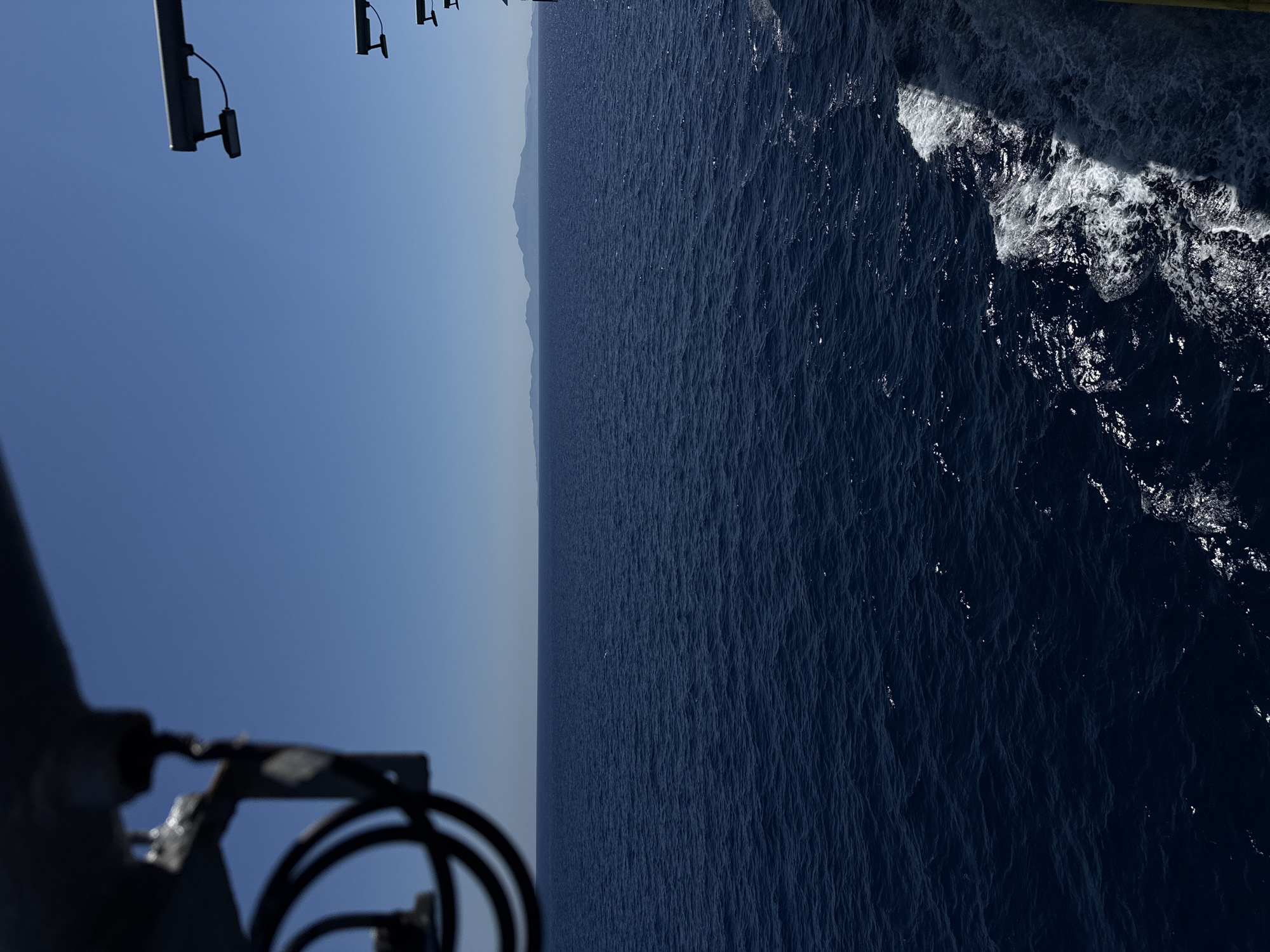 The image shows a view of the sea from a vessel sailing near Kalamia, Greece. The water is a deep blue, with small waves creating a textured surface. In the distance, a hazy outline of land is visible on the horizon under a clear blue sky. The foreground shows part of the vessel, including a metal railing and some equipment with cables. The wake of the boat is visible trailing behind on the right side of the image, with white foam and disturbed water. The scene depicts a calm day at sea.