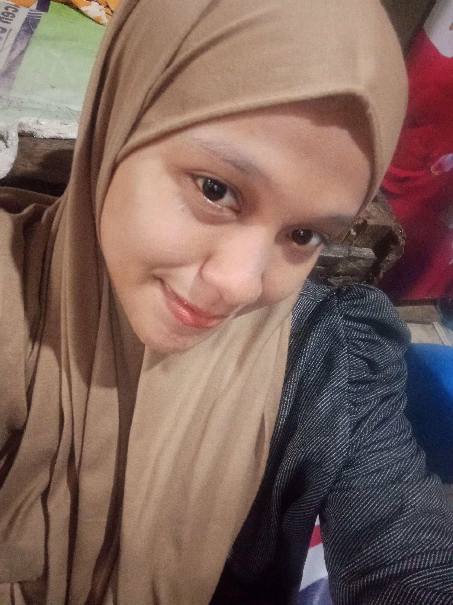 The image is a close-up, selfie-style portrait of a young woman wearing a light brown hijab and a dark jacket with a subtle pinstripe pattern. Her face is turned slightly towards the camera, and she is smiling. Her eyes are dark and her lips are slightly colored. The background is blurred and appears to be indoors, with some indistinct objects and textures visible, possibly indicating a casual or domestic setting. There is no specific information in the image that suggests a location in Meunasahara, Indonesia.