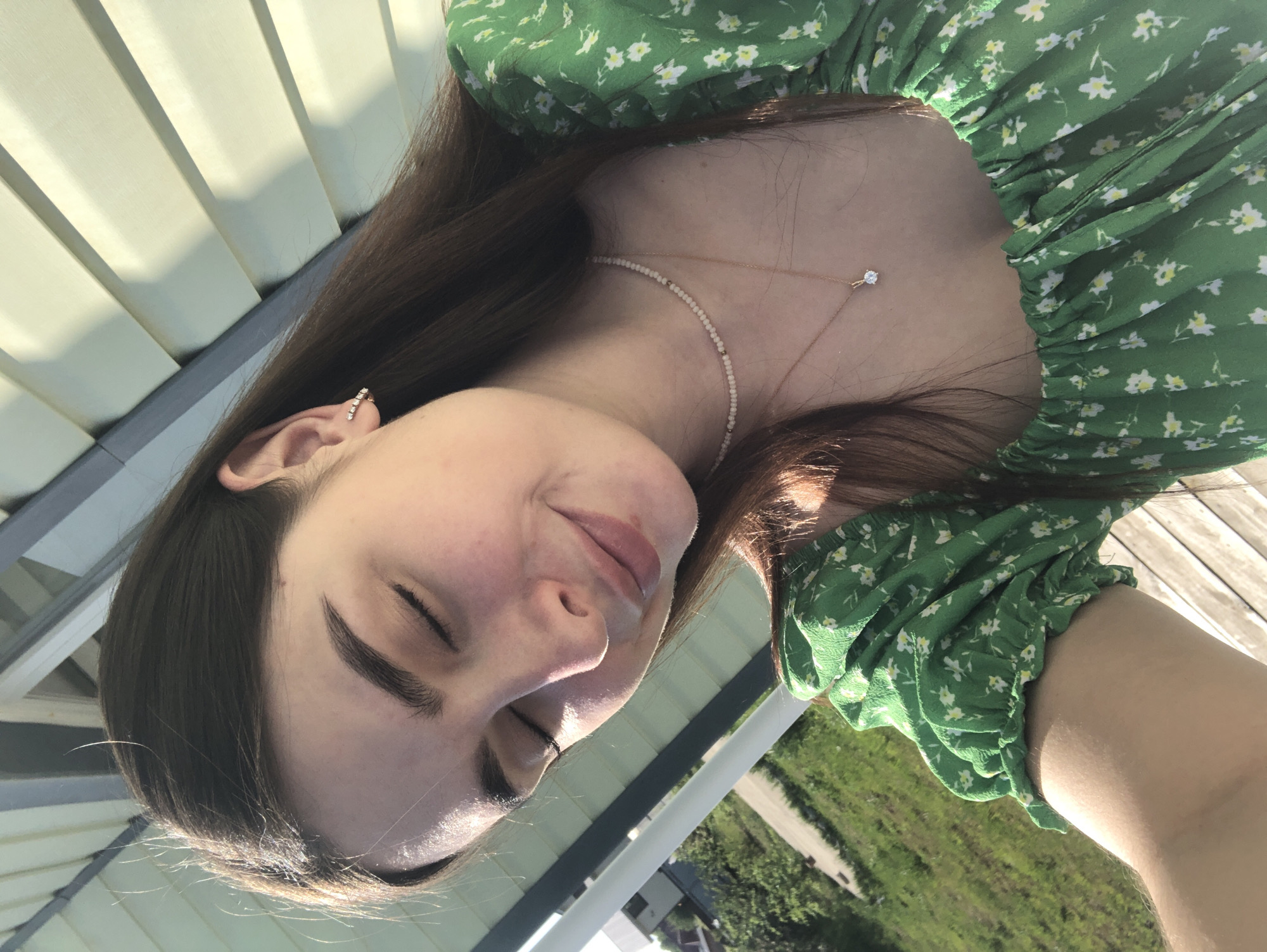 A young woman is angled in the image, with her eyes closed. She is wearing a green floral dress and two necklaces. Her hair is brown and straight. She has an earring on her left ear. The background includes an open outdoor area with grass and some buildings. The image does not contain enough information to determine the location in Noviy, Russia.