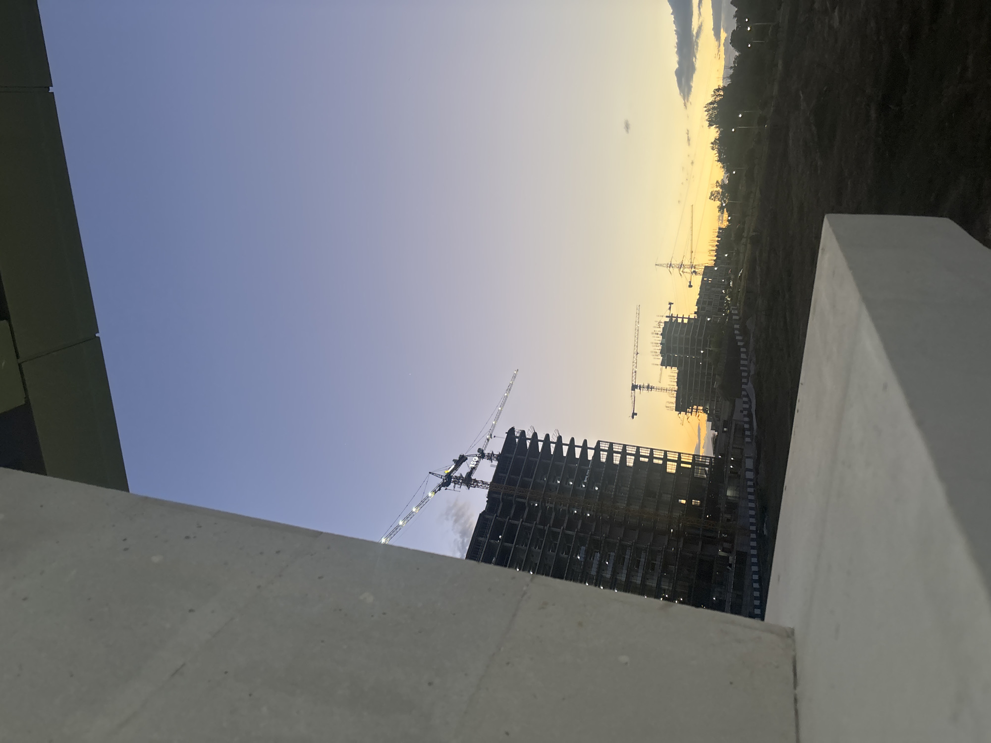 The media file captures an outdoor panoramic view of a large construction site in Yerevan, Armenia, during either sunrise or sunset.
In the foreground, a light-colored, rough concrete wall or barrier spans the bottom of the frame and extends up the right side, partially framing the scene.
The midground is dominated by multiple large, multi-story buildings in various stages of construction. The closest building on the left shows a detailed concrete skeletal structure with numerous floors and openings, suggesting ongoing development. Several tall construction cranes are positioned around these structures, their booms extending high into the sky, some with lights illuminated. These cranes signify active building work.
In the background, a natural landscape unfolds with distant hills or mountains covered in dark vegetation, stretching towards the horizon. Faint utility poles and power lines are discernible against the glowing sky.
The sky is a prominent feature, displaying a striking gradient of colors characteristic of dawn or dusk. It transitions from a clear, deep blue at the top of the frame to a warm, vibrant yellow and orange glow along the horizon, casting a soft, ambient light over the entire scene. A few indistinct clouds are visible near the brighter horizon.
The overall image conveys a sense of ongoing urban development and construction activity. No people or visible text are present in the image.